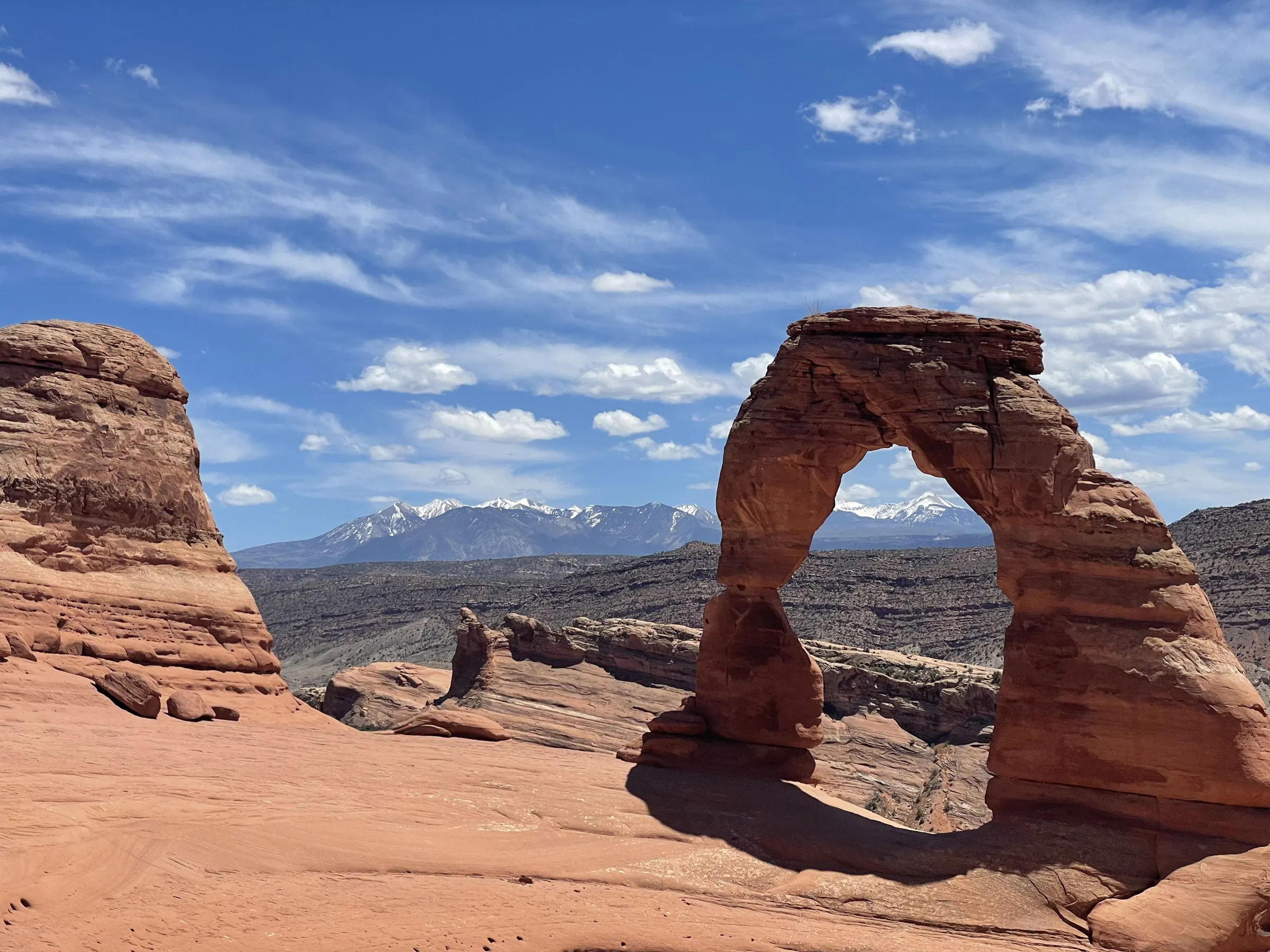 Delicate Arch, a free-standing curve of red sandstone, on a bare rock bowl with snow-covered mountains on the horizon