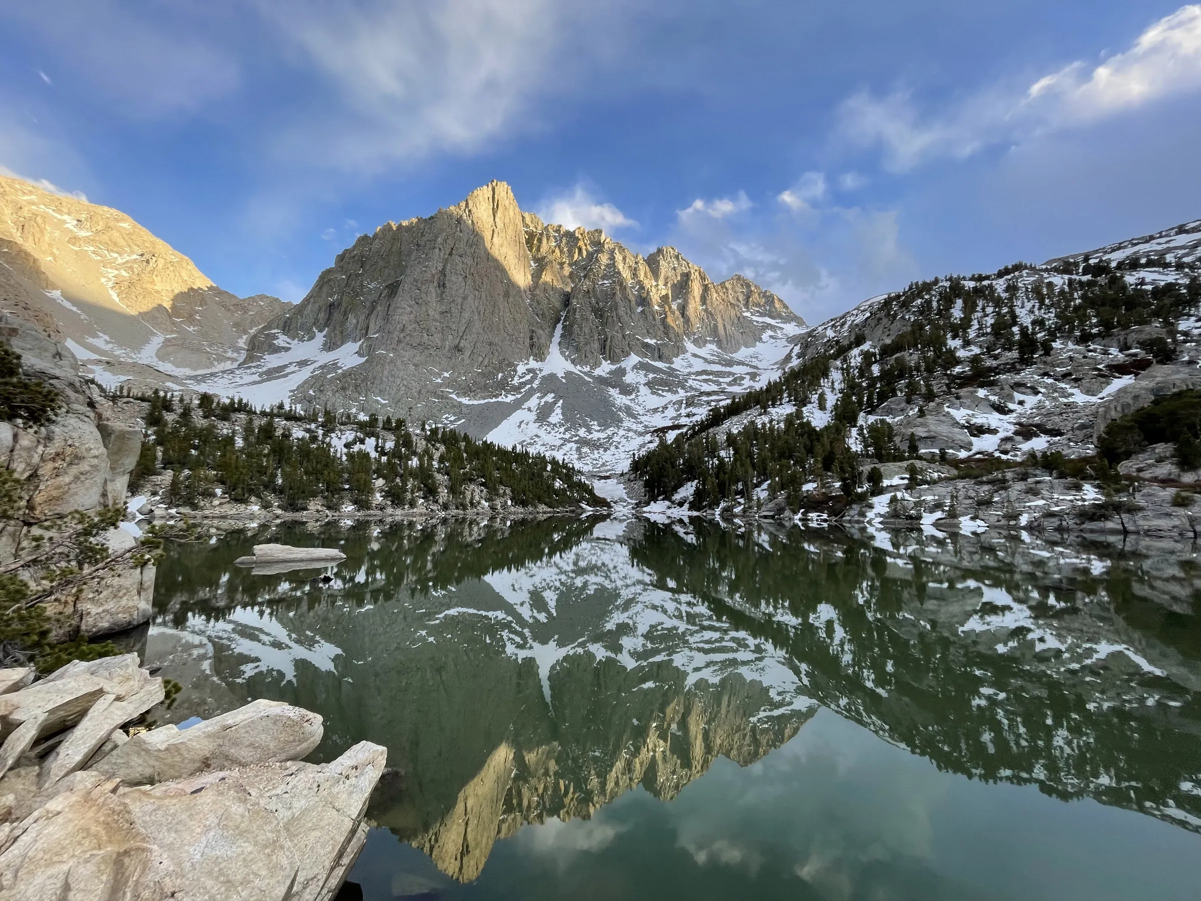 A jagged granite peak catching early sun above a still alpine lake, with snow on the slopes and pines along the shore