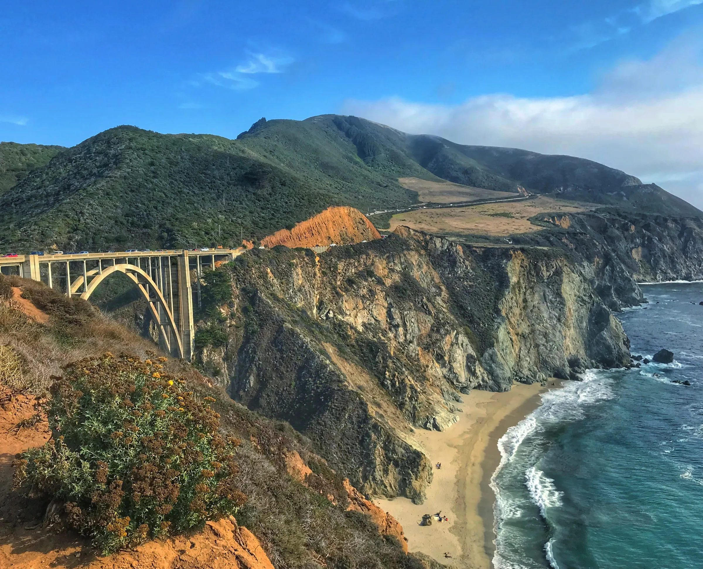 A concrete arch bridge spanning a canyon on a steep green coastline, with fog spilling over the ridge and surf on the beach below