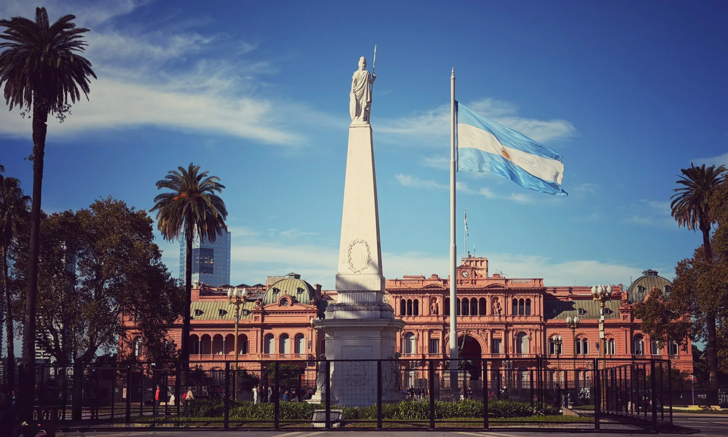 A long pink presidential palace behind a tall white monument topped by a robed figure holding a staff, with the Argentine flag flying from a pole beside it and palms on either side