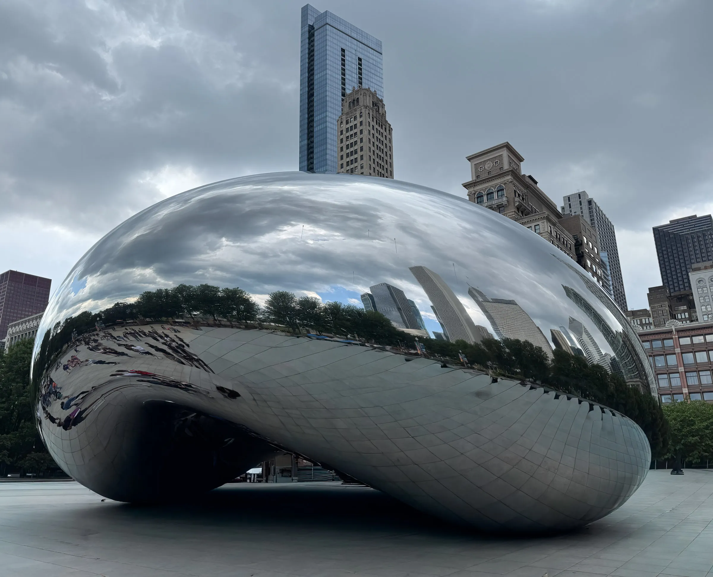 The mirrored steel Cloud Gate sculpture on a plaza, its curved surface reflecting a stretched skyline and the people beneath it, with towers and heavy grey cloud behind