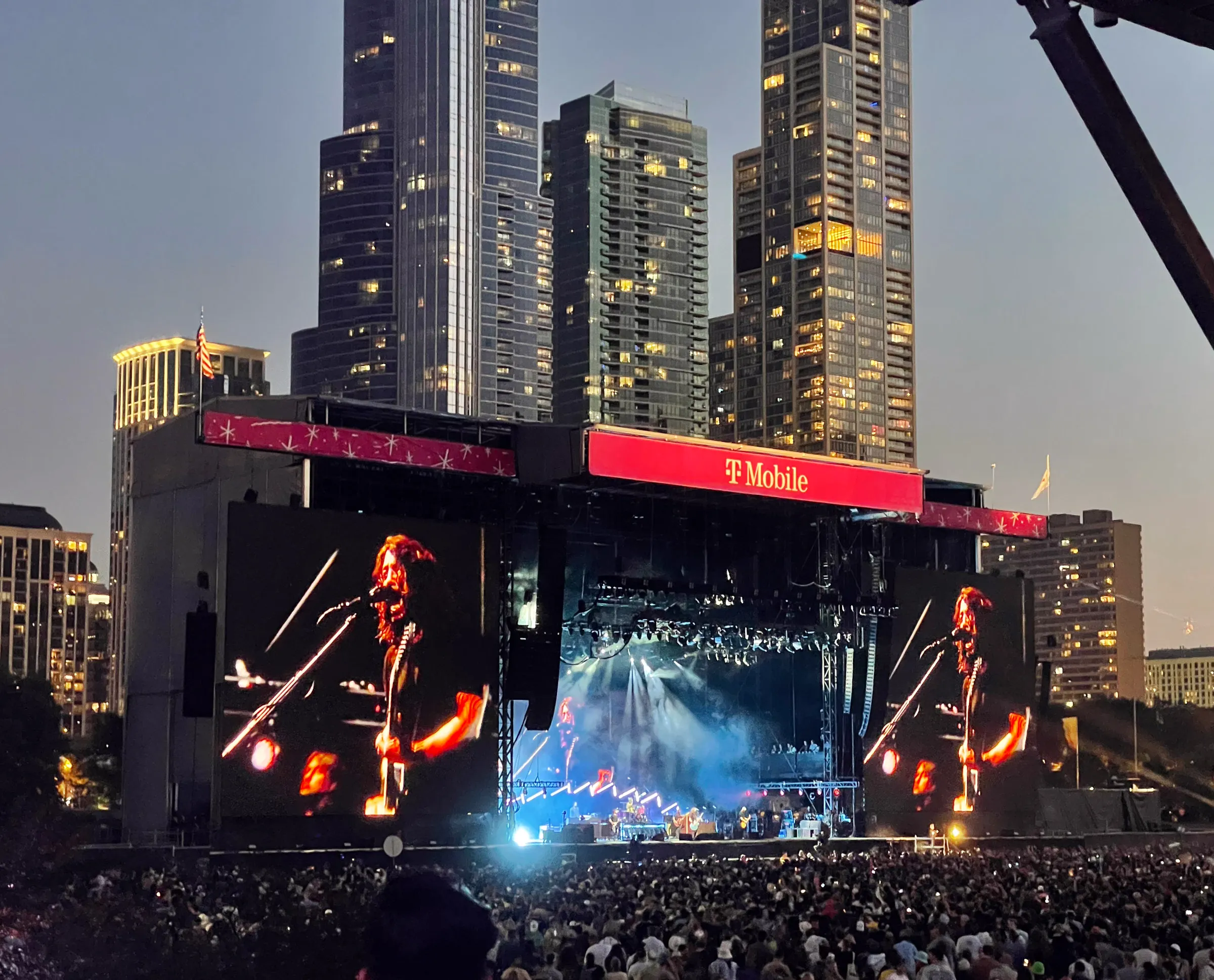 A festival crowd filling a park at dusk in front of a large stage, the band small under the lights and a guitarist shown huge on the screens to either side, glass towers rising behind
