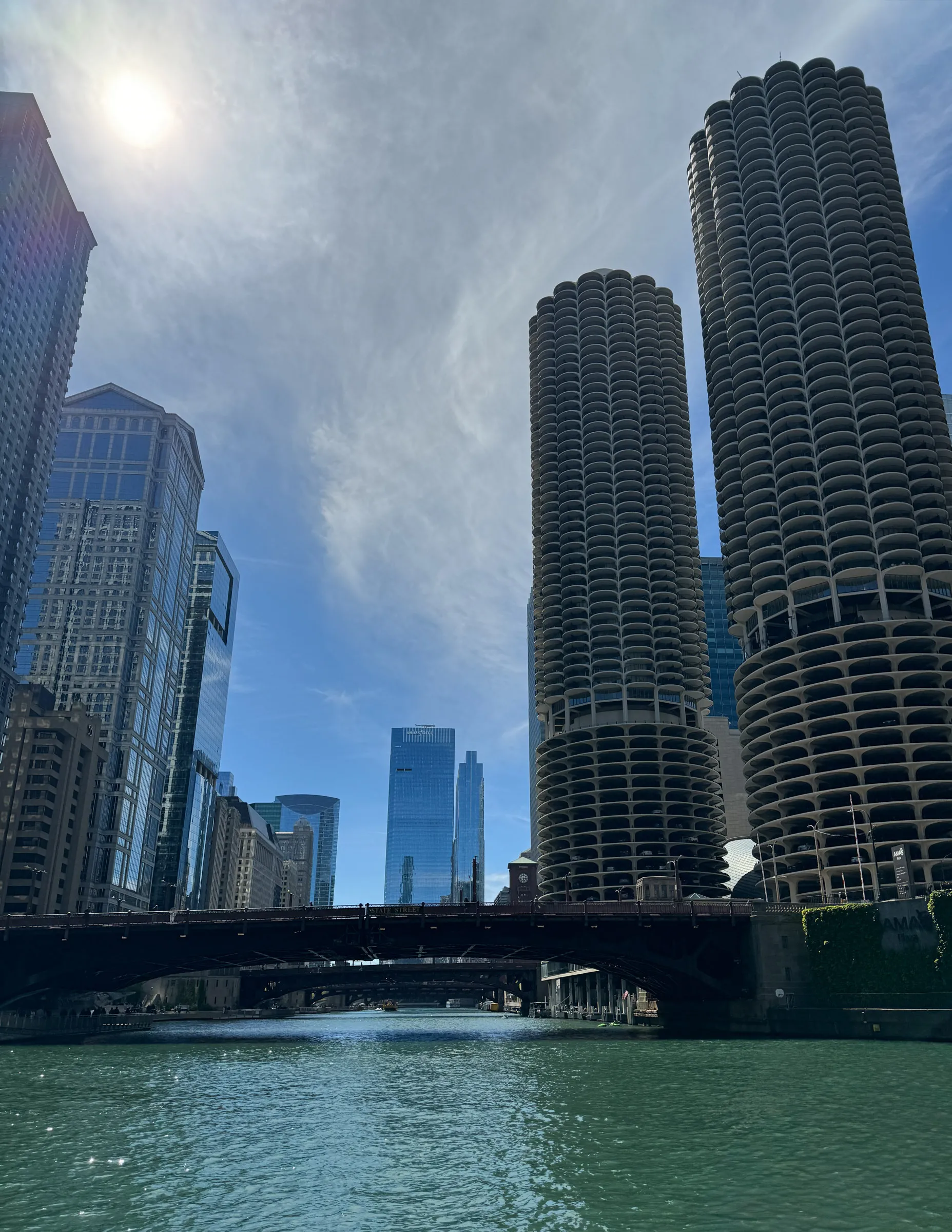 Looking up the Chicago River from water level at the two round corncob towers of Marina City, with a bridge crossing ahead and the sun flaring over the buildings to the left