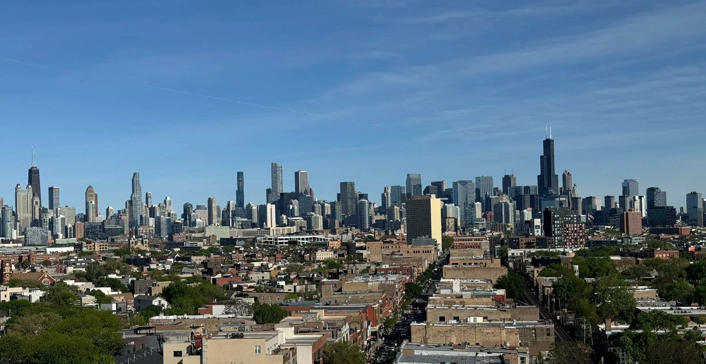 The Chicago skyline standing on the horizon beyond low rooftops and treetops, its two tallest towers at either end of the cluster under a clear blue sky