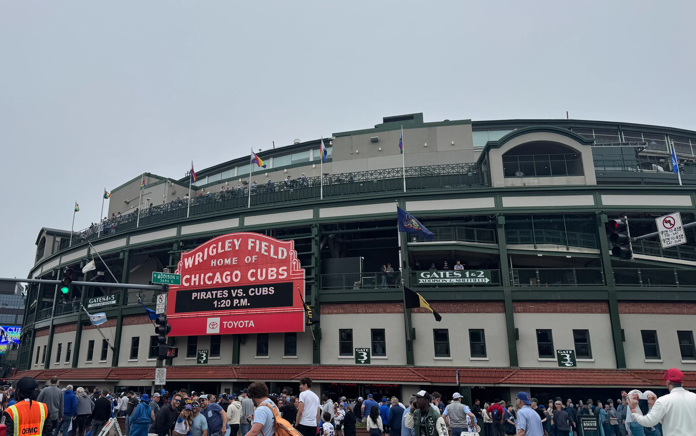 The red Wrigley Field marquee reading Home of the Chicago Cubs above a board announcing Pirates vs Cubs at 1:20 pm, with crowds filling the sidewalk under the ballpark's green facade