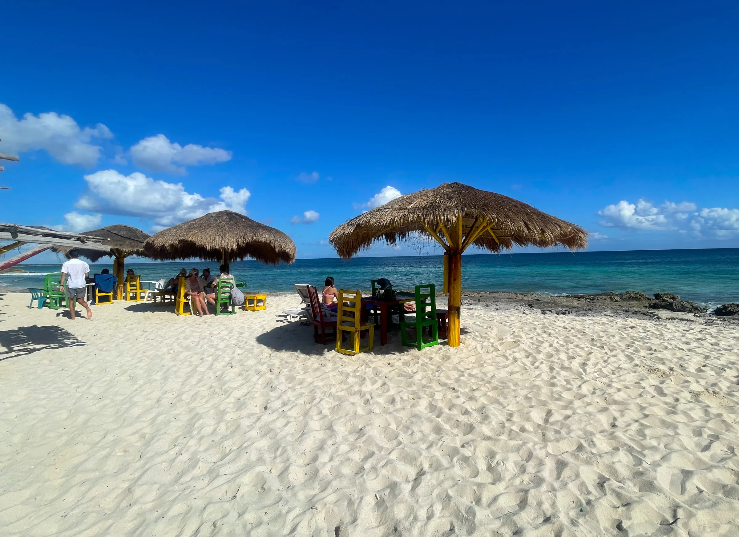 Thatched palapas and brightly painted wooden chairs on white sand, a few people sitting in the shade, turquoise water beyond