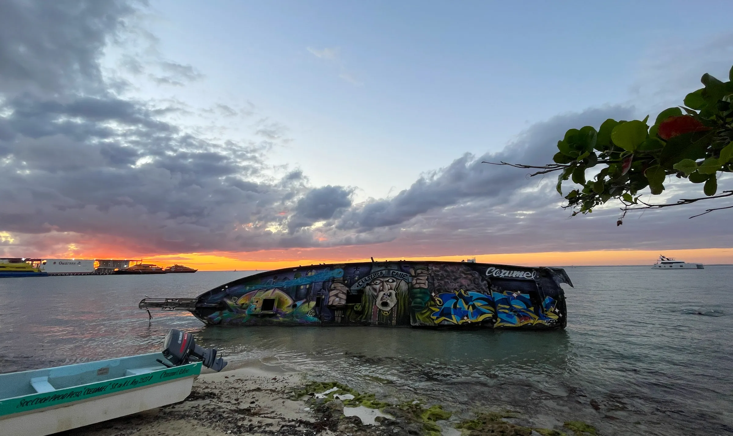 A wrecked boat hull covered end to end in graffiti, lying half in the shallows at dusk, with an orange sunset behind it and a small blue skiff on the sand