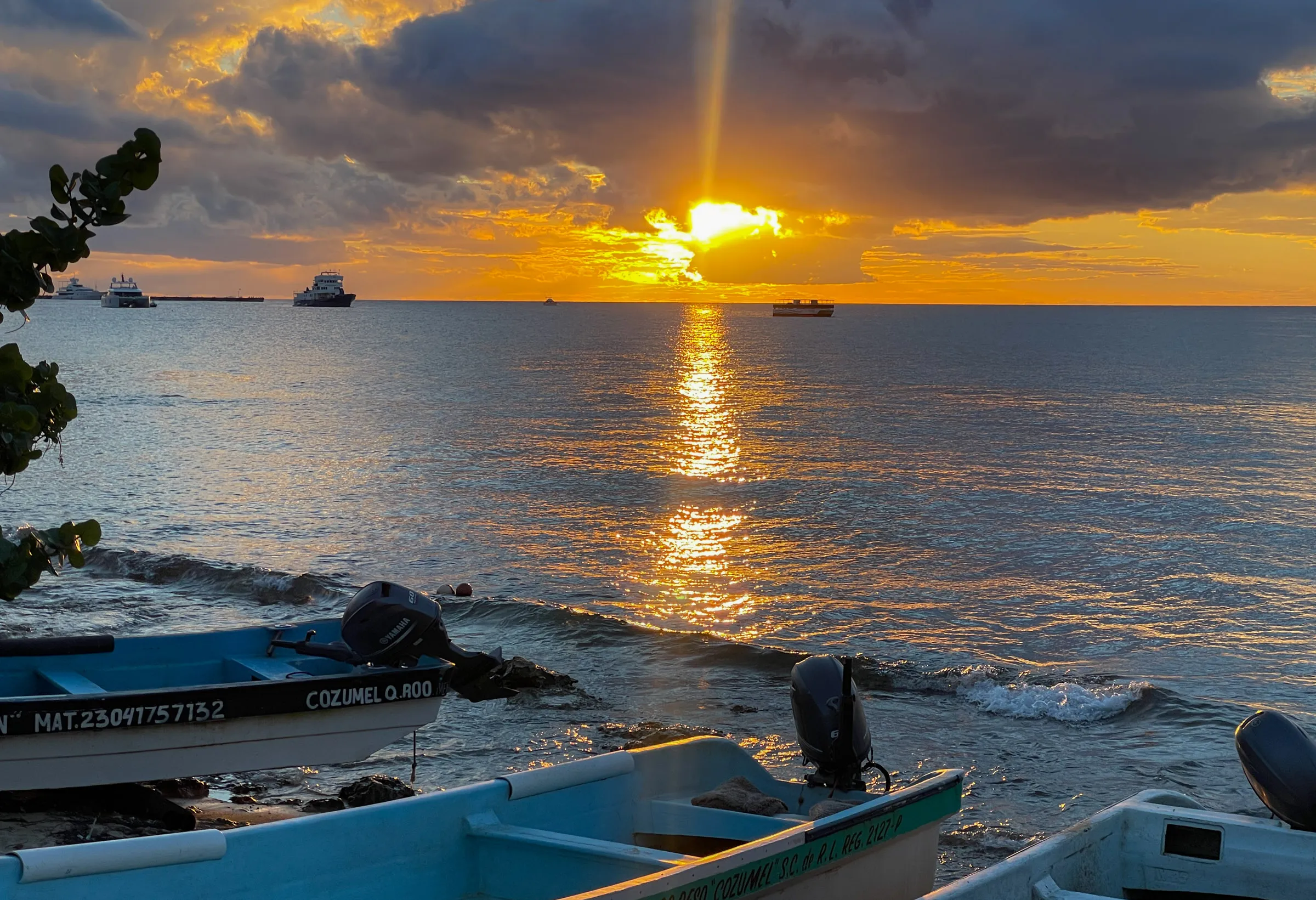 The sun dropping through heavy cloud over the sea and laying a bright track across the water, with small outboard boats pulled up on the rocks in the foreground