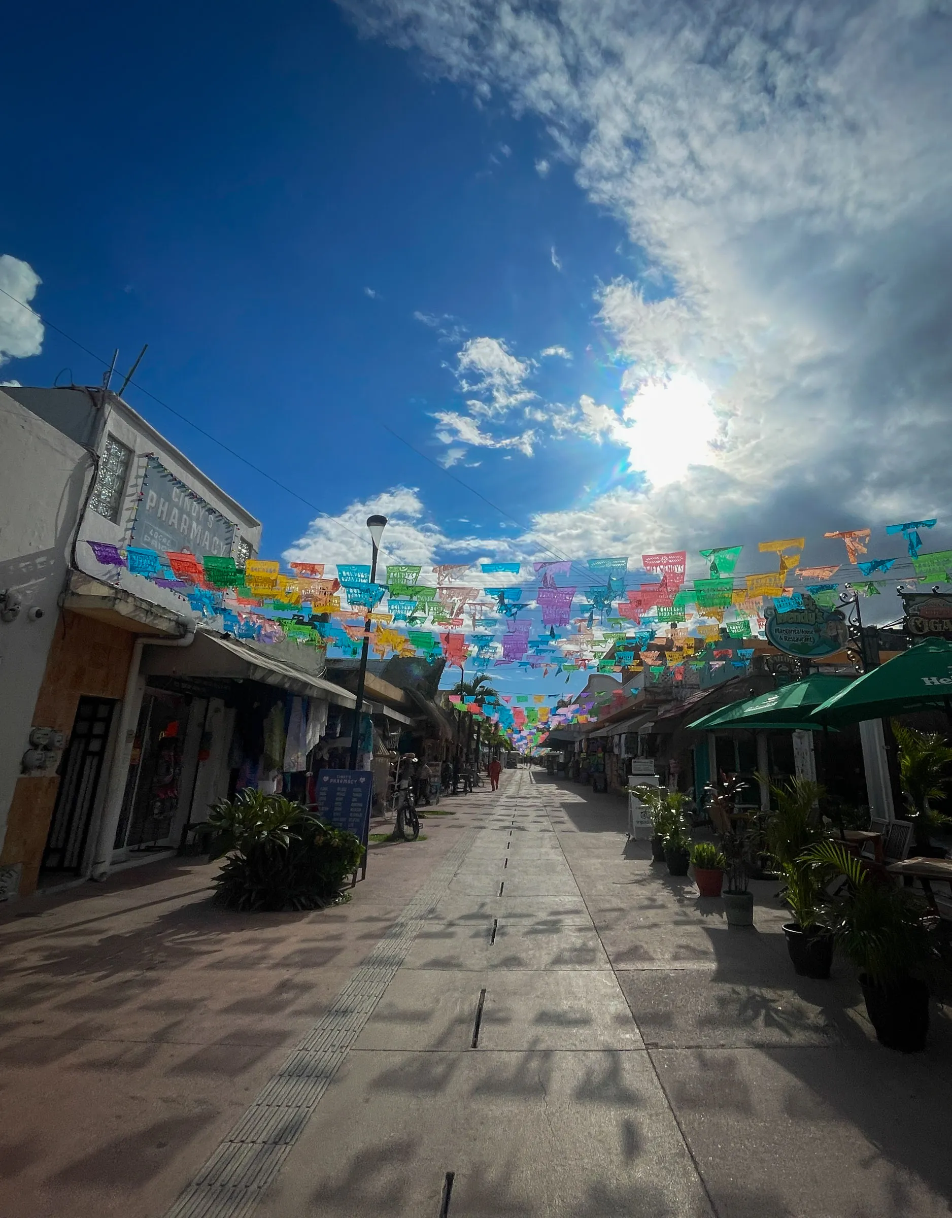 A pedestrian street of low shopfronts with rows of colourful papel picado strung overhead, the sun breaking through cloud above