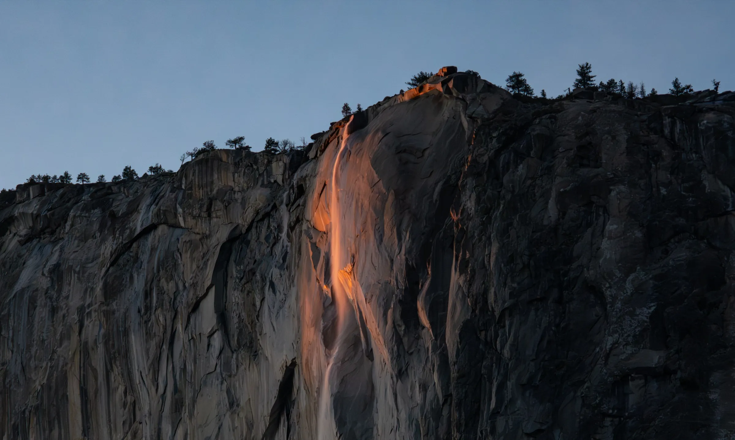 A narrow waterfall on a dark granite cliff, lit bright orange by the setting sun against a pale blue sky
