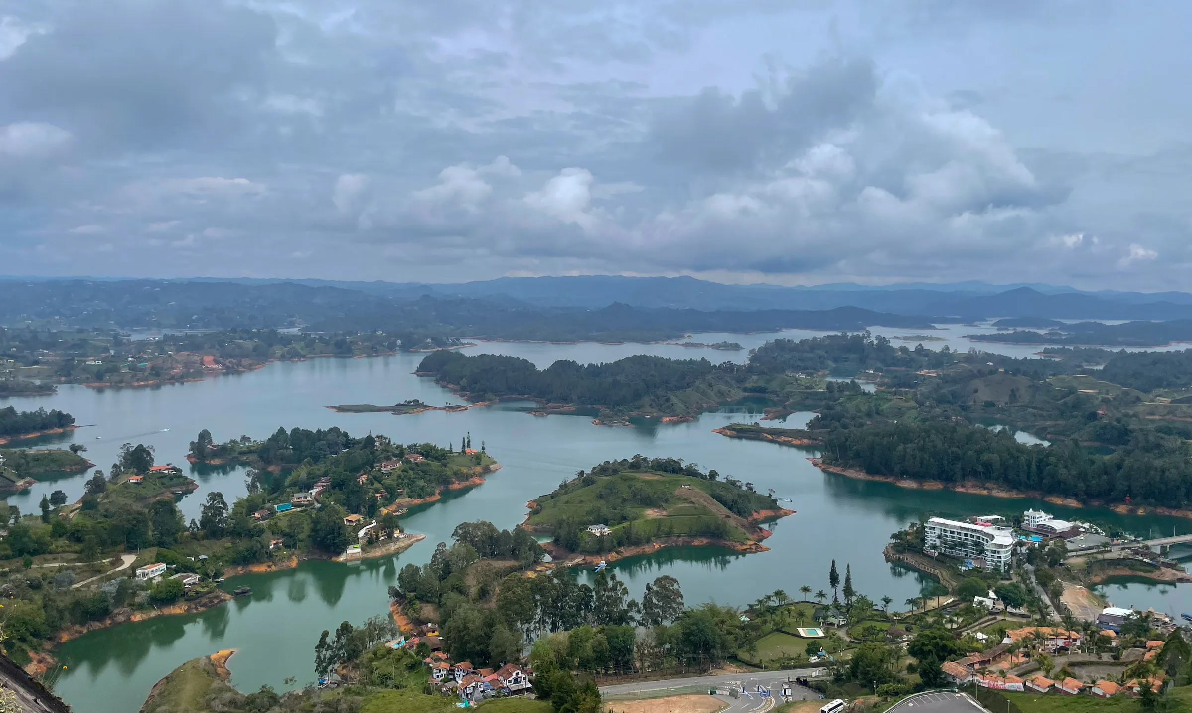 A high view over a green reservoir winding between wooded islands and headlands, with hazy ridges on the horizon under heavy cloud