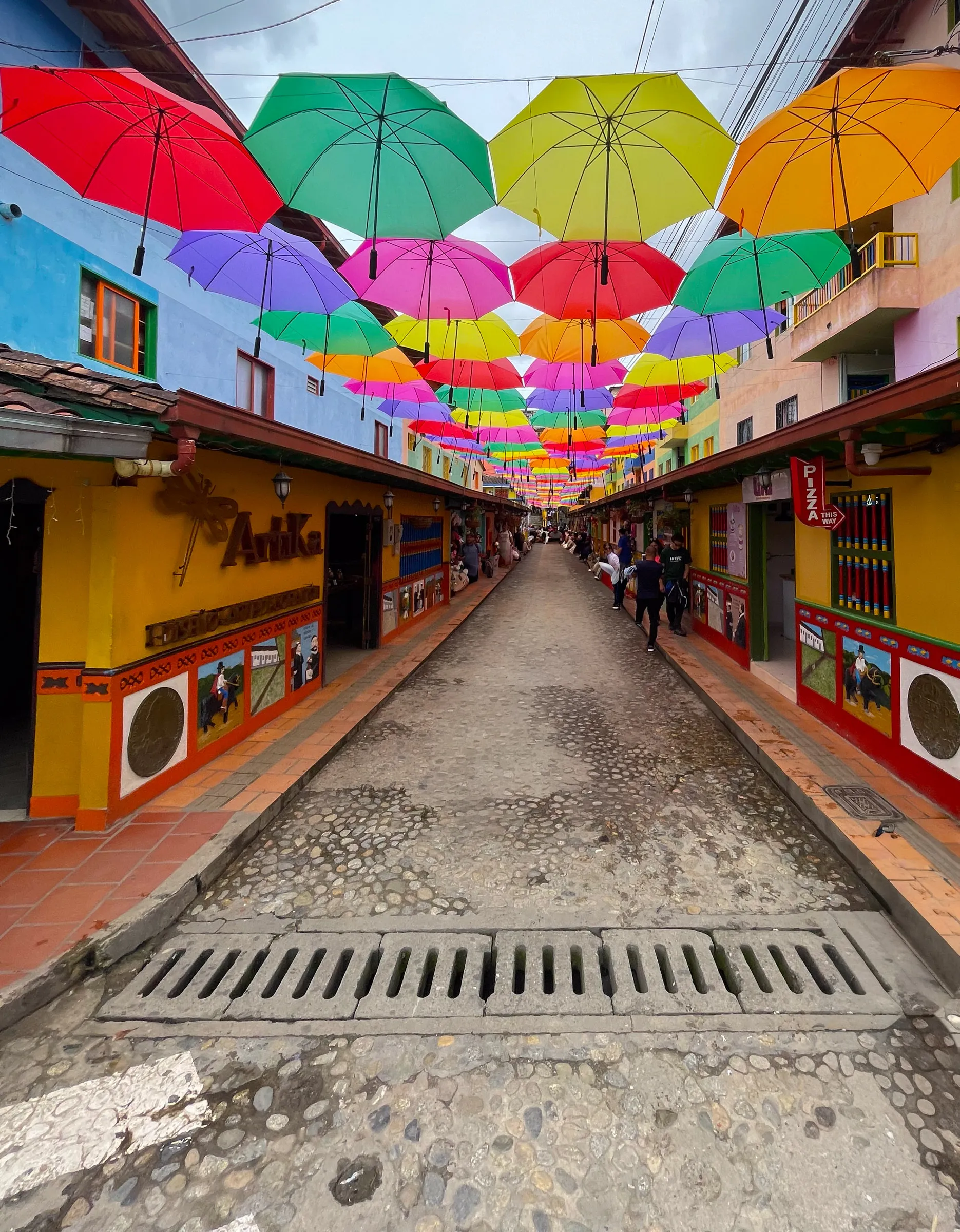 A narrow cobbled street lined with buildings painted yellow, red and blue, with rows of open umbrellas in every colour strung overhead