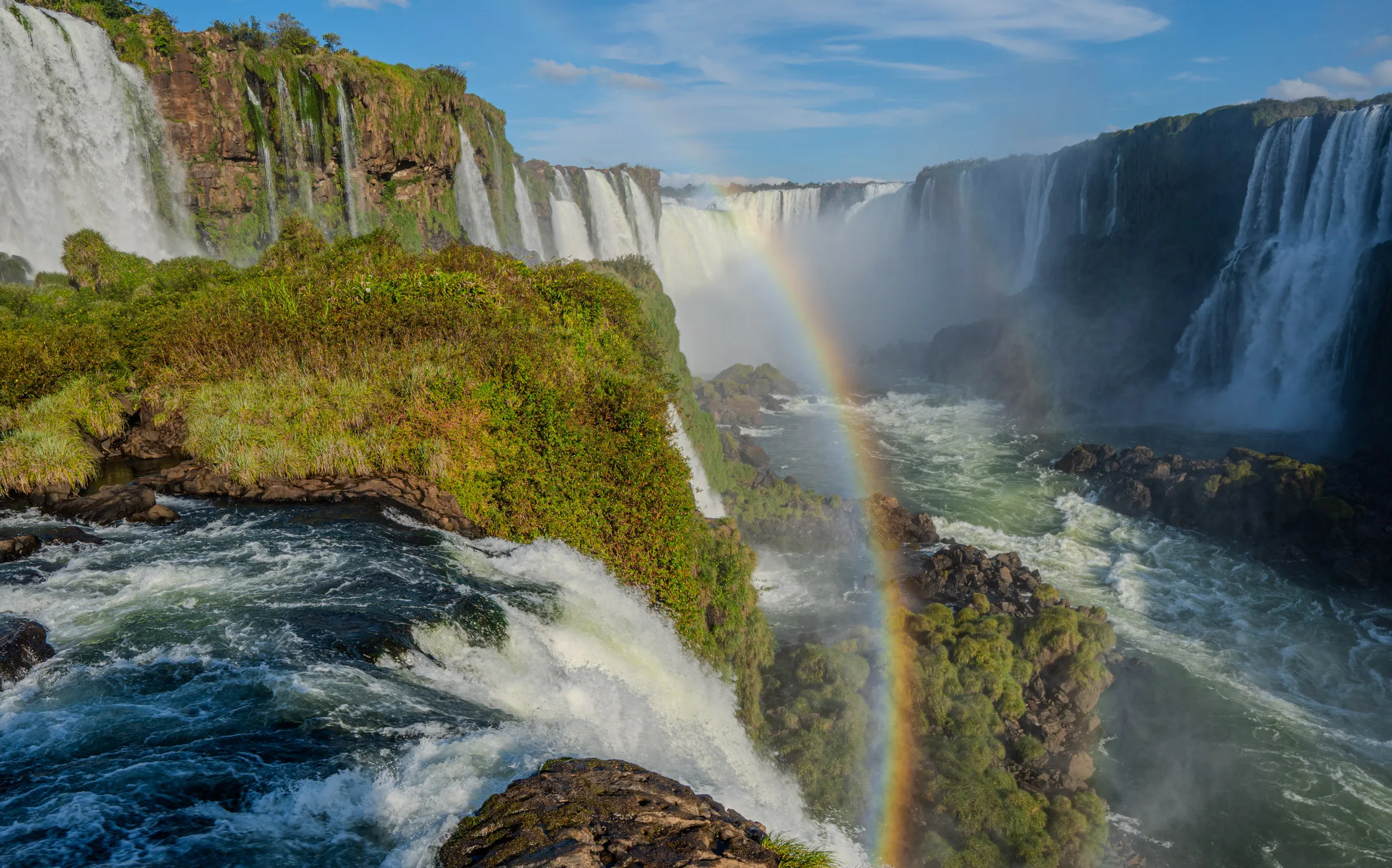 A wide curtain of waterfalls dropping between green cliffs into a river gorge, with a rainbow standing in the mist