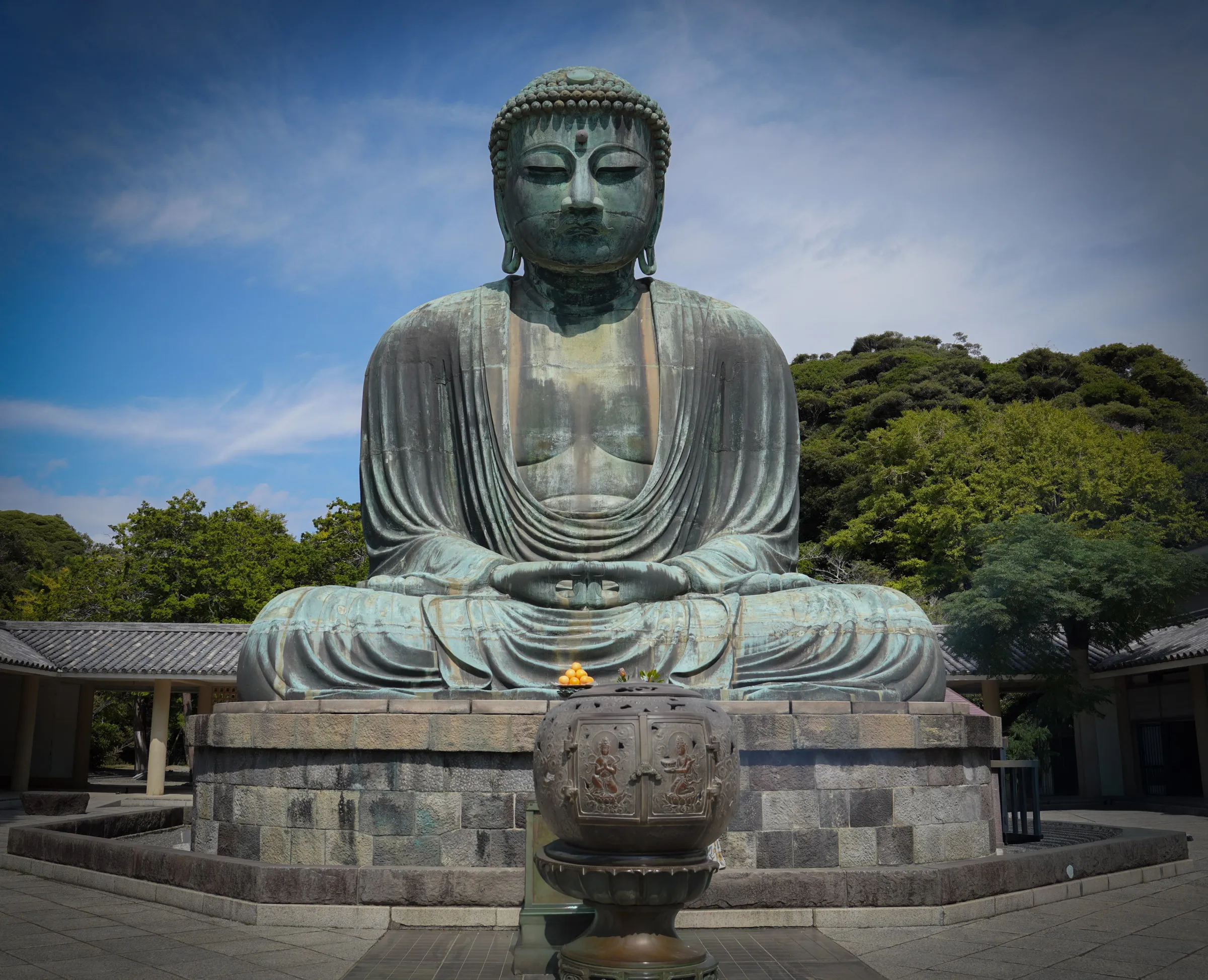 A large bronze seated Buddha statue with a weathered green patina, hands folded in its lap, an offering of oranges set before it and forested hills behind