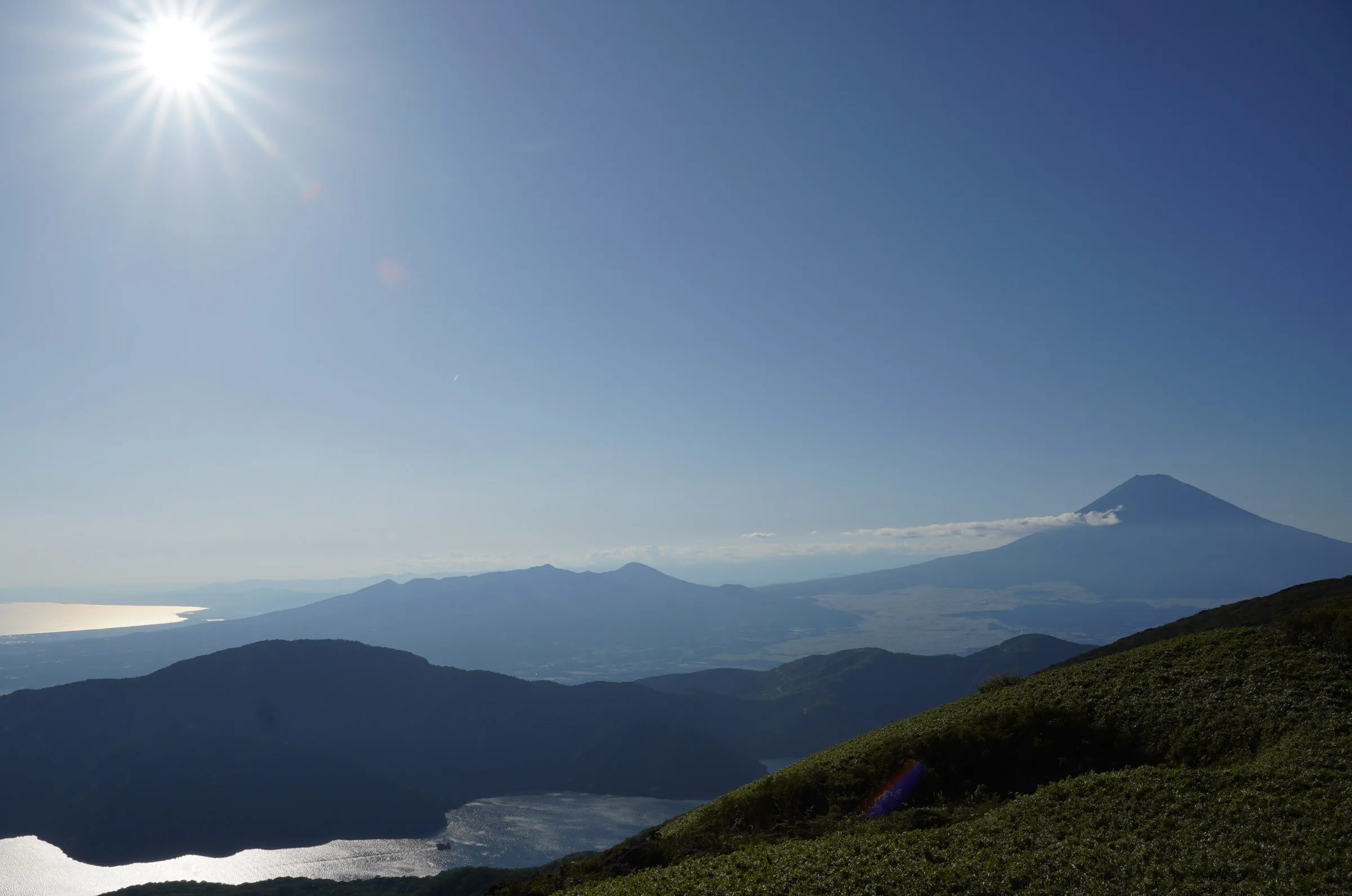 A hazy view from a mountain ridge over Lake Ashi and layered hills to the silhouette of Mount Fuji on the horizon, with the sun flaring at the top of the frame