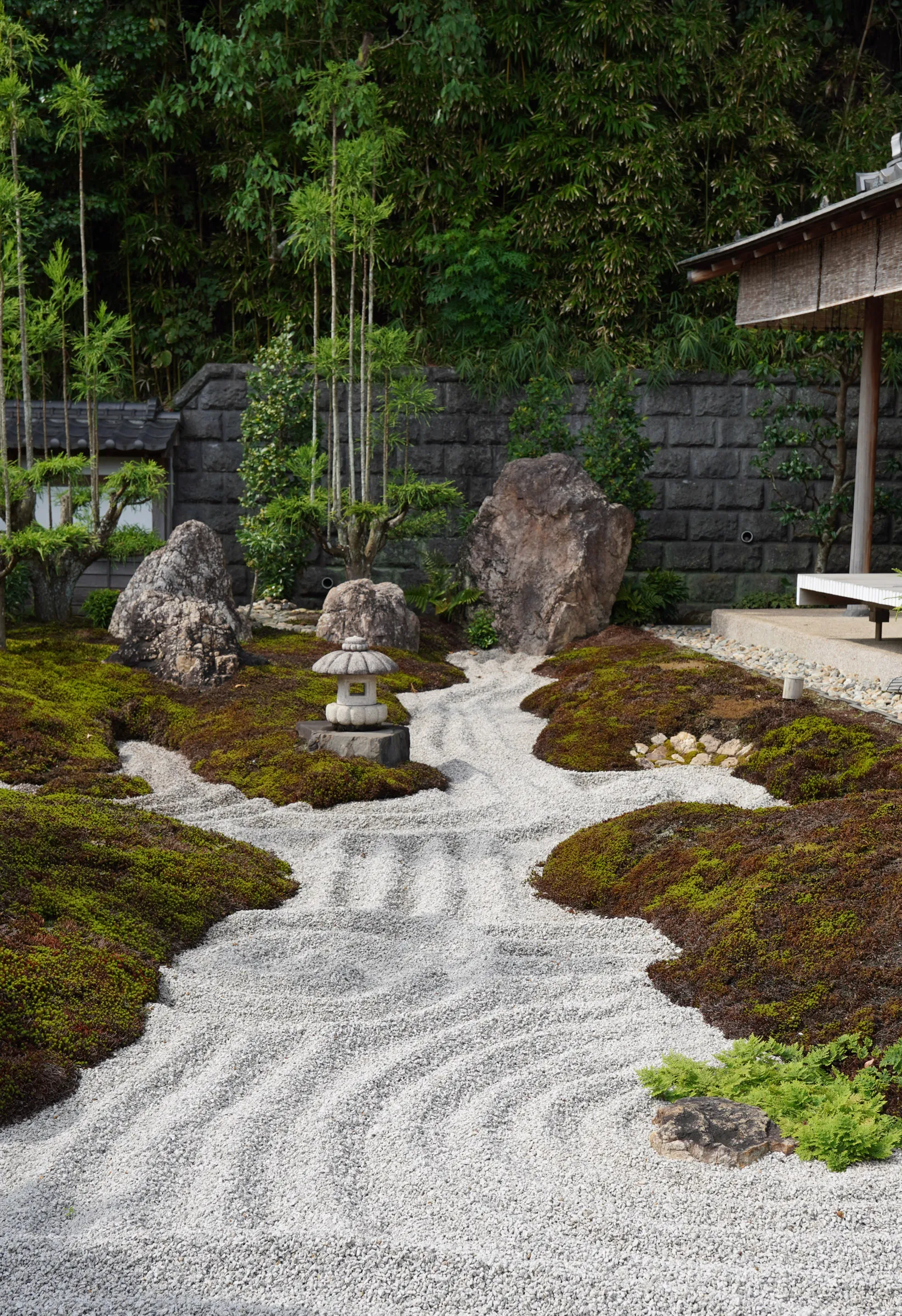 A raked white gravel Zen garden winding between mounds of moss and large upright rocks, with a stone lantern and a bamboo grove behind a stone wall