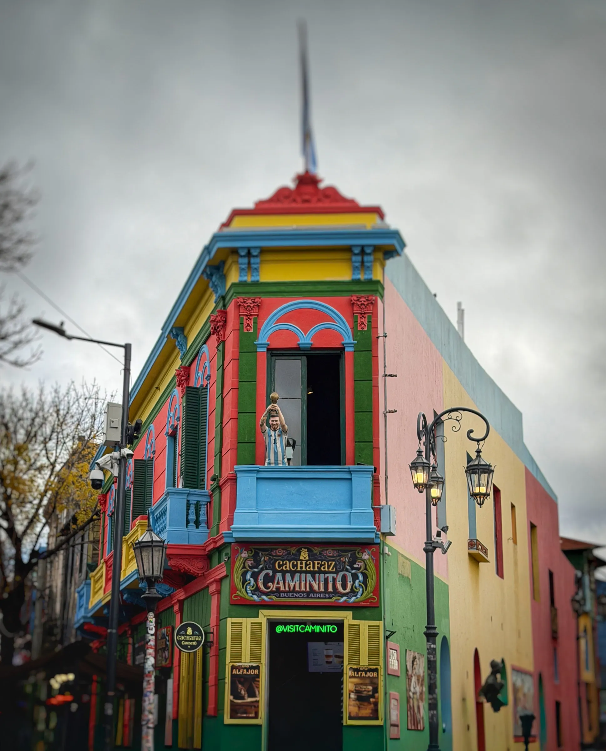A corner building painted in blocks of red, yellow, blue and green, with a cutout of a footballer in an Argentina shirt holding a trophy standing on its balcony under a grey sky