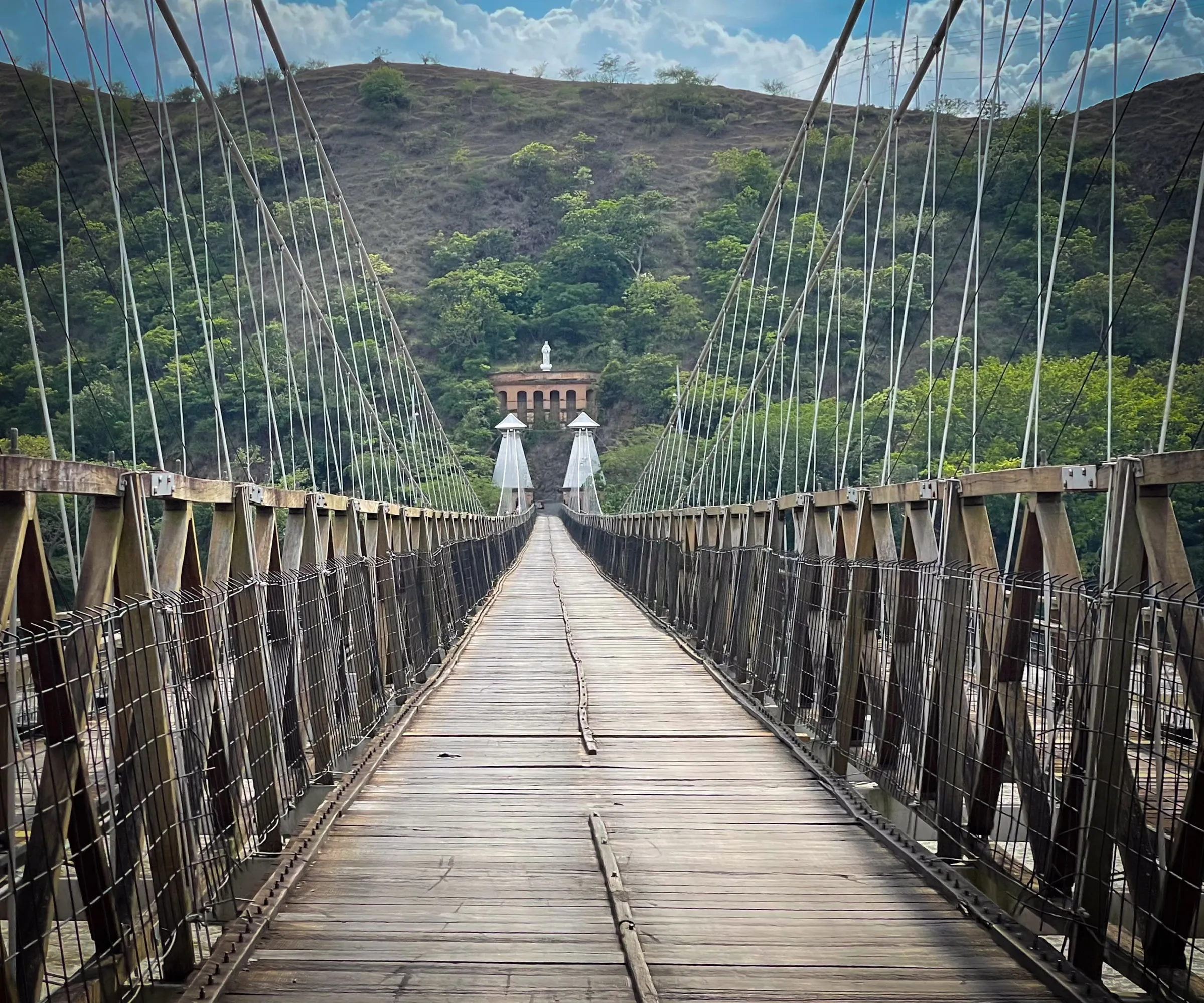 A long suspension bridge with a plank deck running straight away from the camera toward a dry green hillside, with a small stone building at the far end