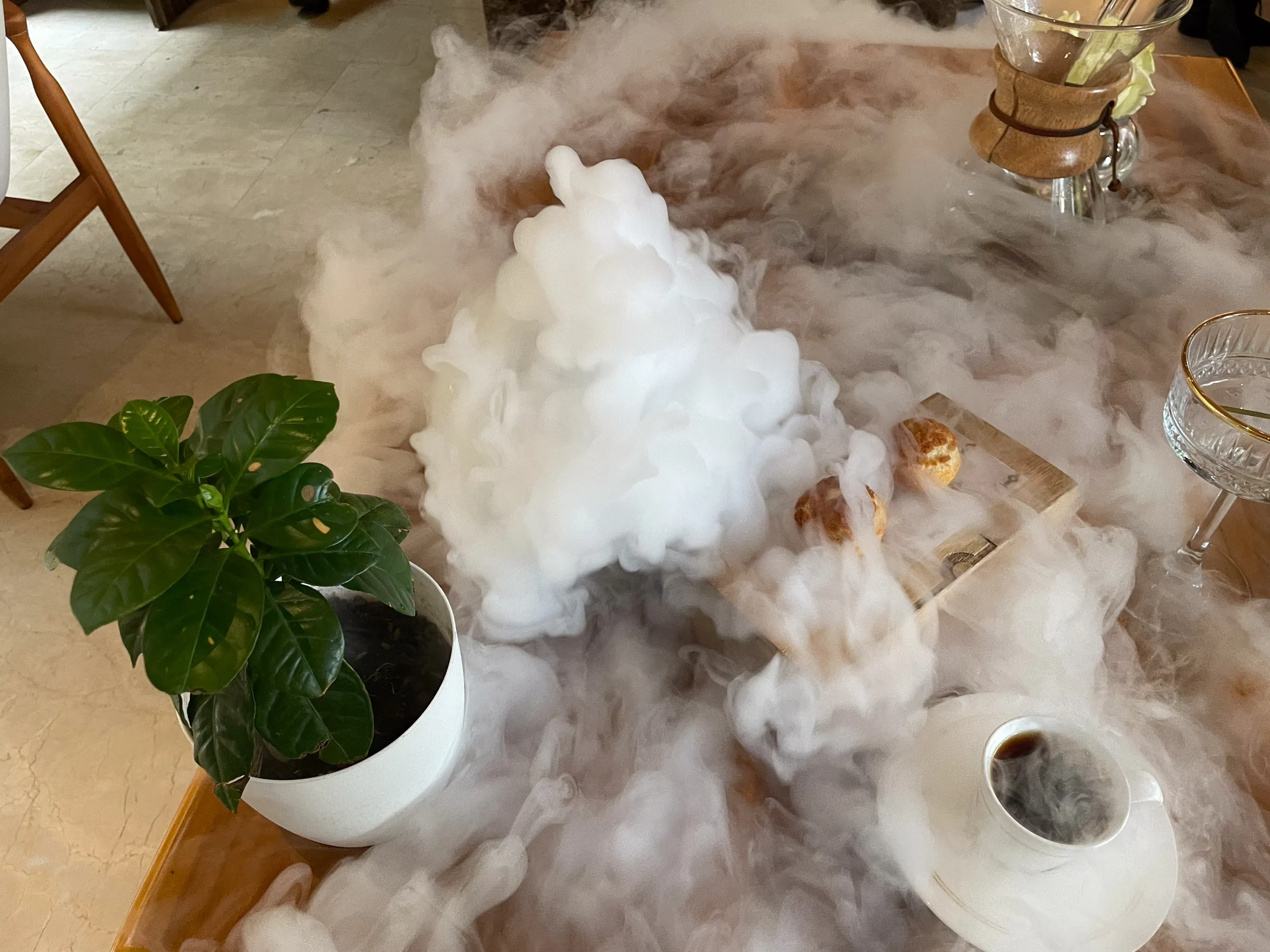 Thick white fog from dry ice pouring across a wooden table set with a Chemex, a cup of coffee, a potted coffee plant and a board of pastries