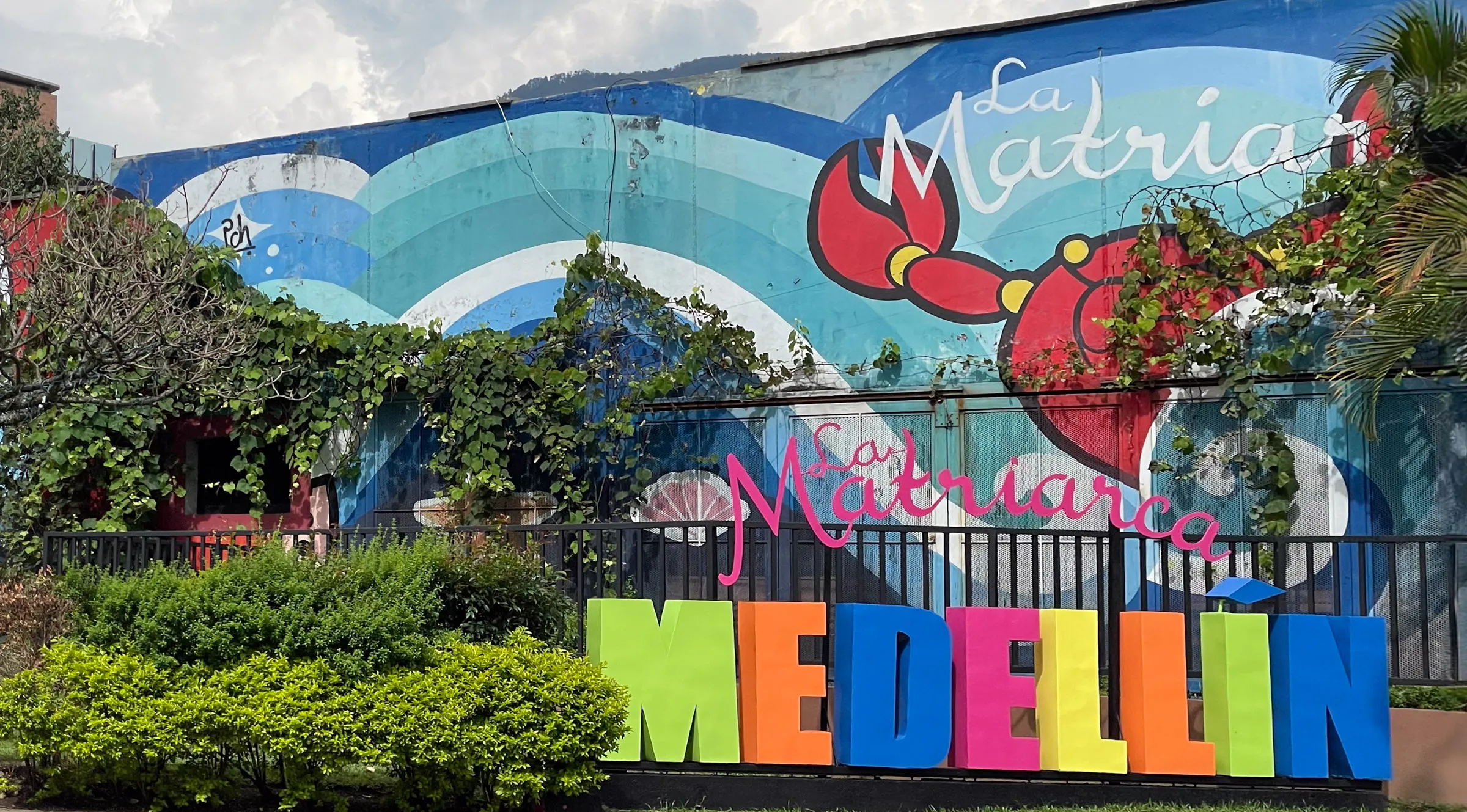 Tall block letters spelling MEDELLÍN in bright colours standing on a lawn, in front of a wall painted with blue waves, a red flower and the words La Matriarca