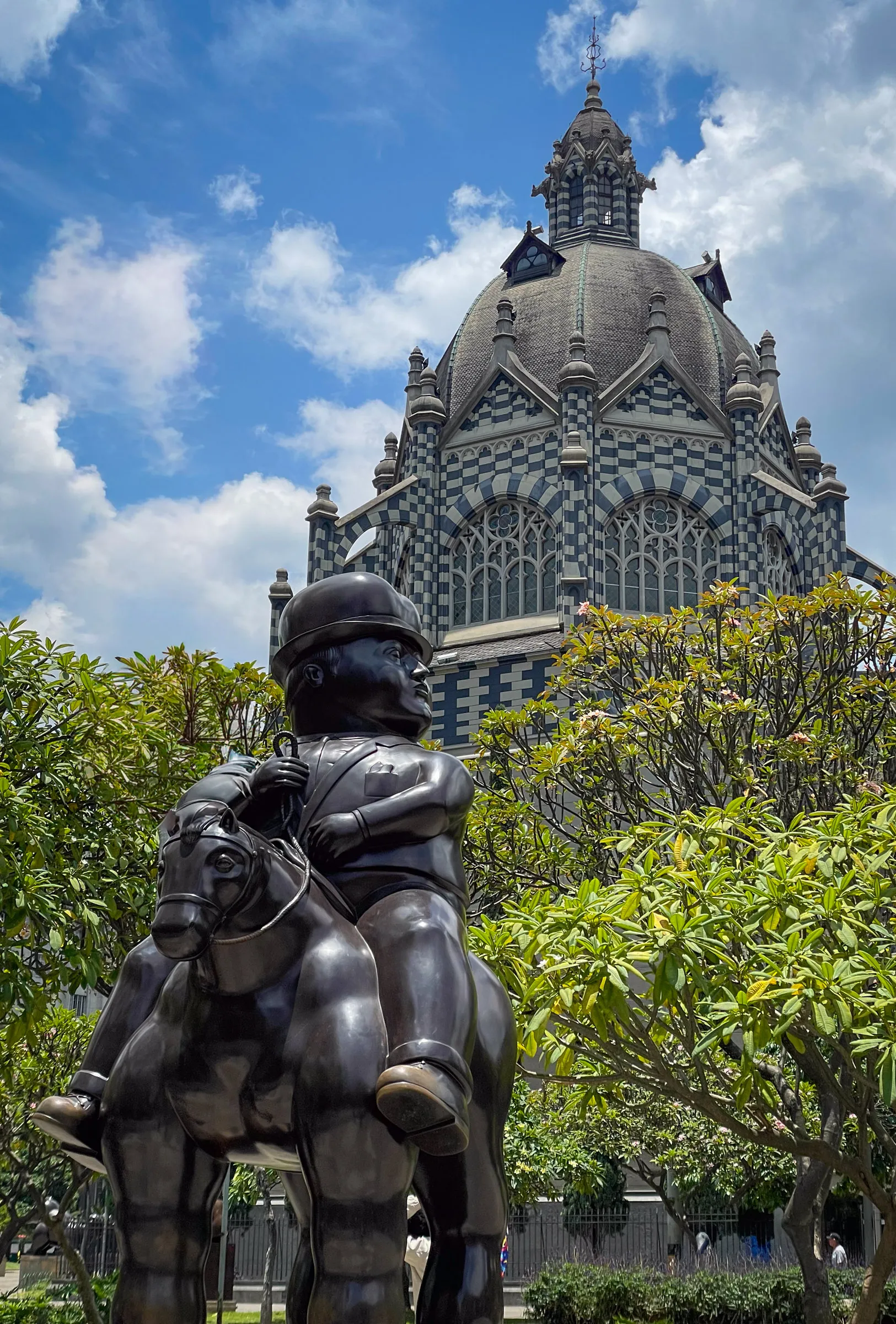 A rounded bronze Botero statue of a helmeted rider on a stout horse, standing among flowering trees in front of the black-and-white patterned dome of the Palacio de la Cultura
