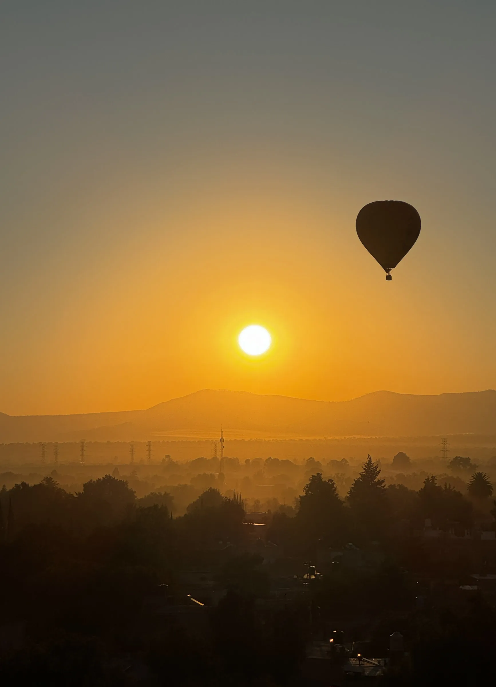 A single hot air balloon hanging high over a hazy valley at sunrise, with the sun clear of a low mountain ridge and trees and rooftops in silhouette below