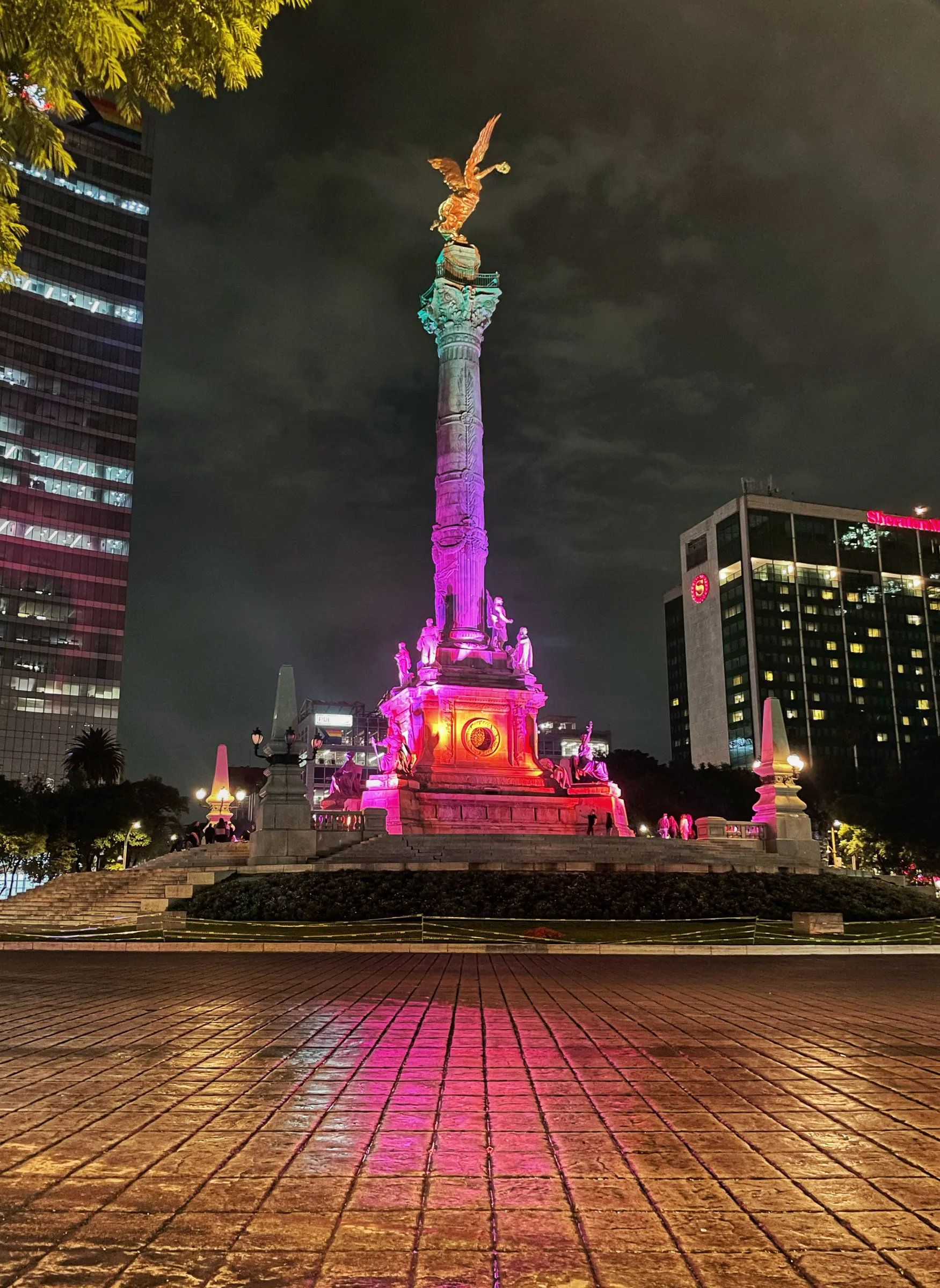 The Angel of Independence column lit pink and gold against a black night sky, the winged gilt figure on top and the wet plaza reflecting the colour back