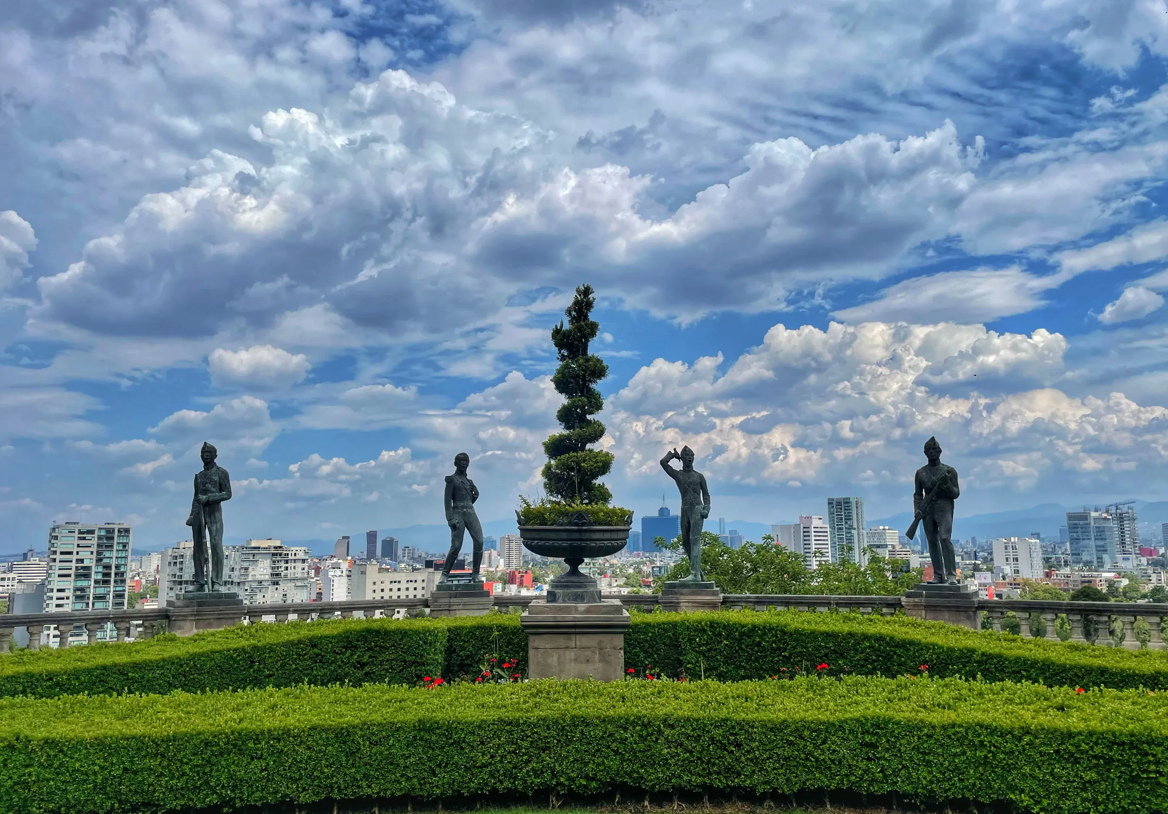 Four bronze statues and a clipped topiary in a stone urn along a castle terrace, with a trimmed hedge in front and the city spreading out under tall cumulus behind