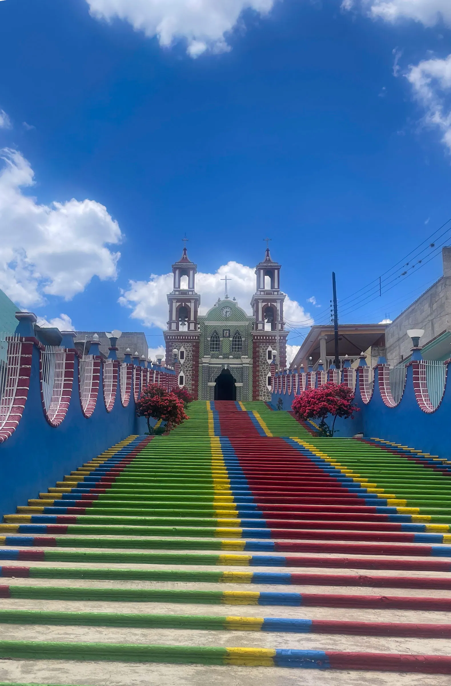 A wide staircase painted in red, yellow, green and blue stripes climbing between blue walls to a twin-towered church under a bright cloudy sky