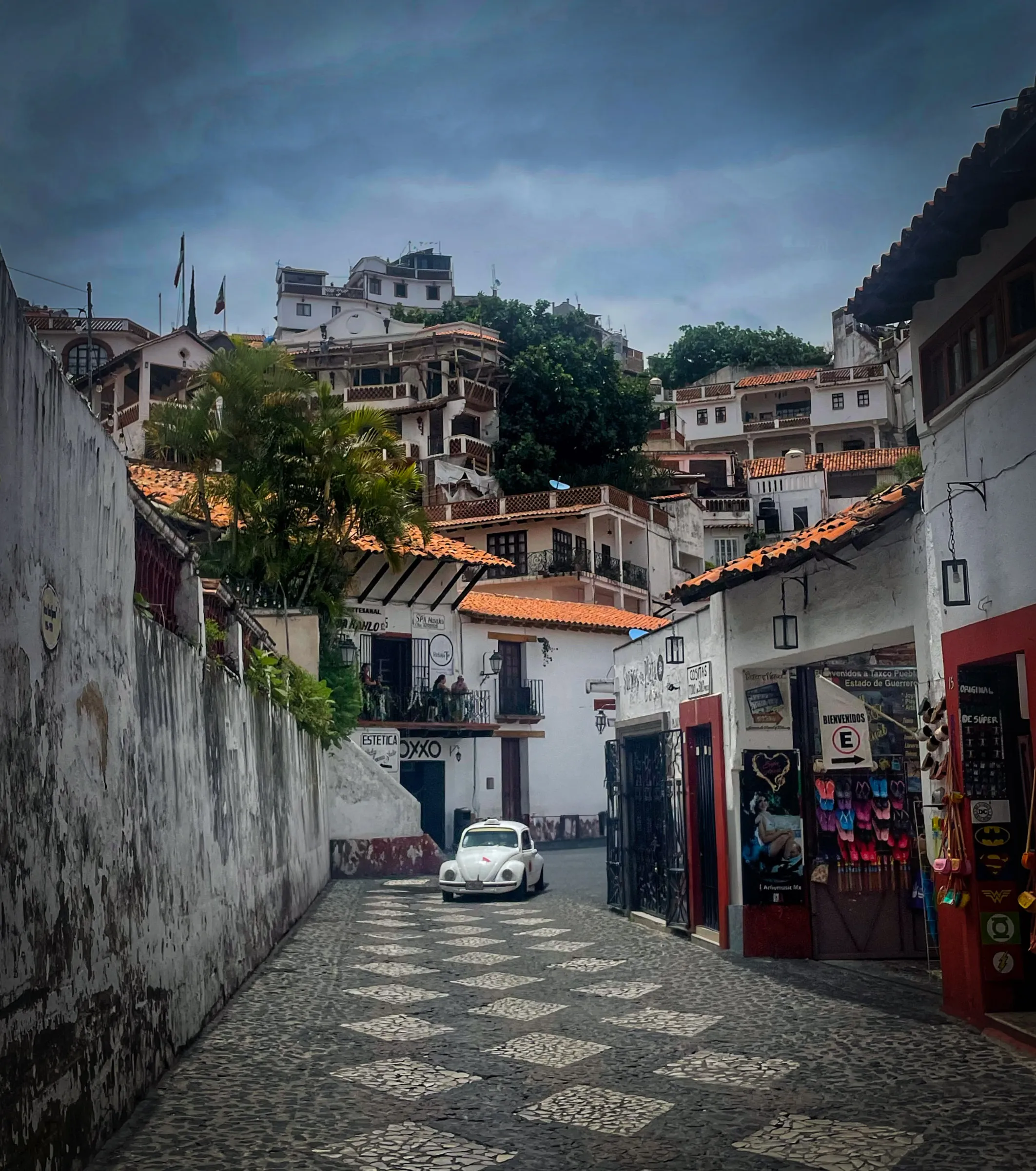 A narrow cobbled street of white buildings with red tiled roofs climbing a steep hillside, a white Volkswagen Beetle parked halfway up under a heavy grey sky