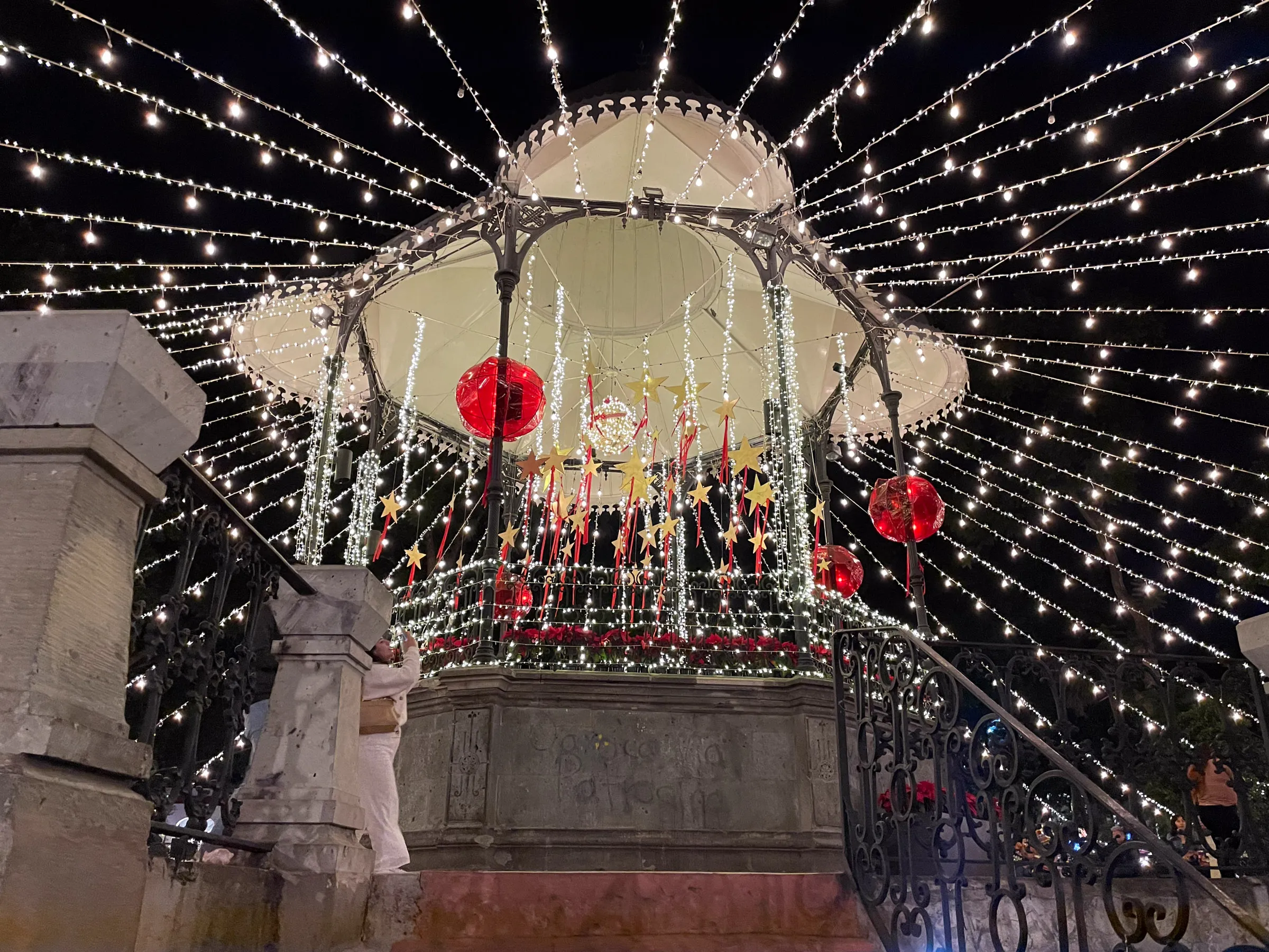 A bandstand in a plaza at night, hung with red baubles and gold stars and wrapped in strings of white lights that fan out overhead in every direction