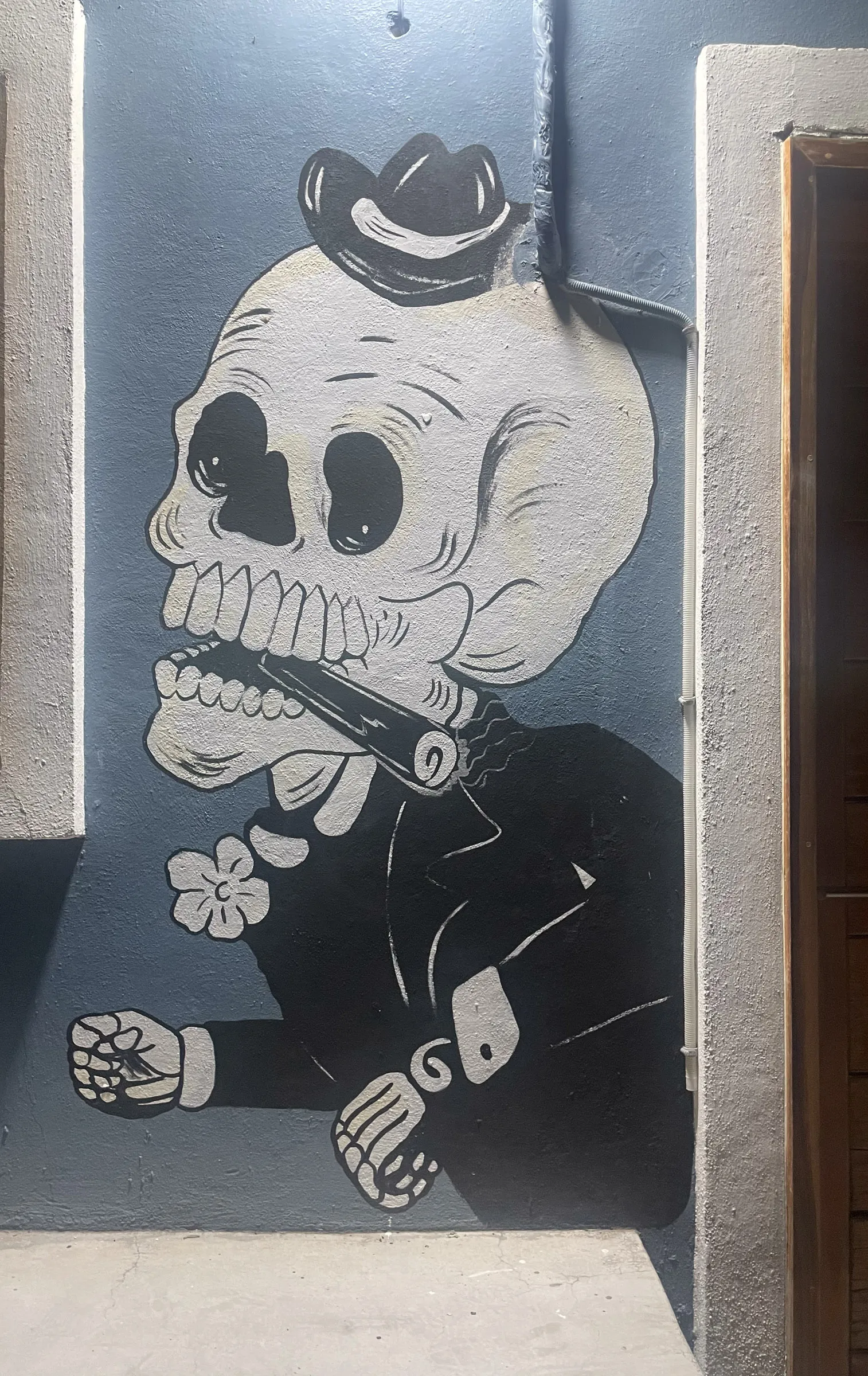 A black and white mural painted in an alcove of a grey wall, showing a skull in a hat and suit smoking a pipe with a flower in one hand