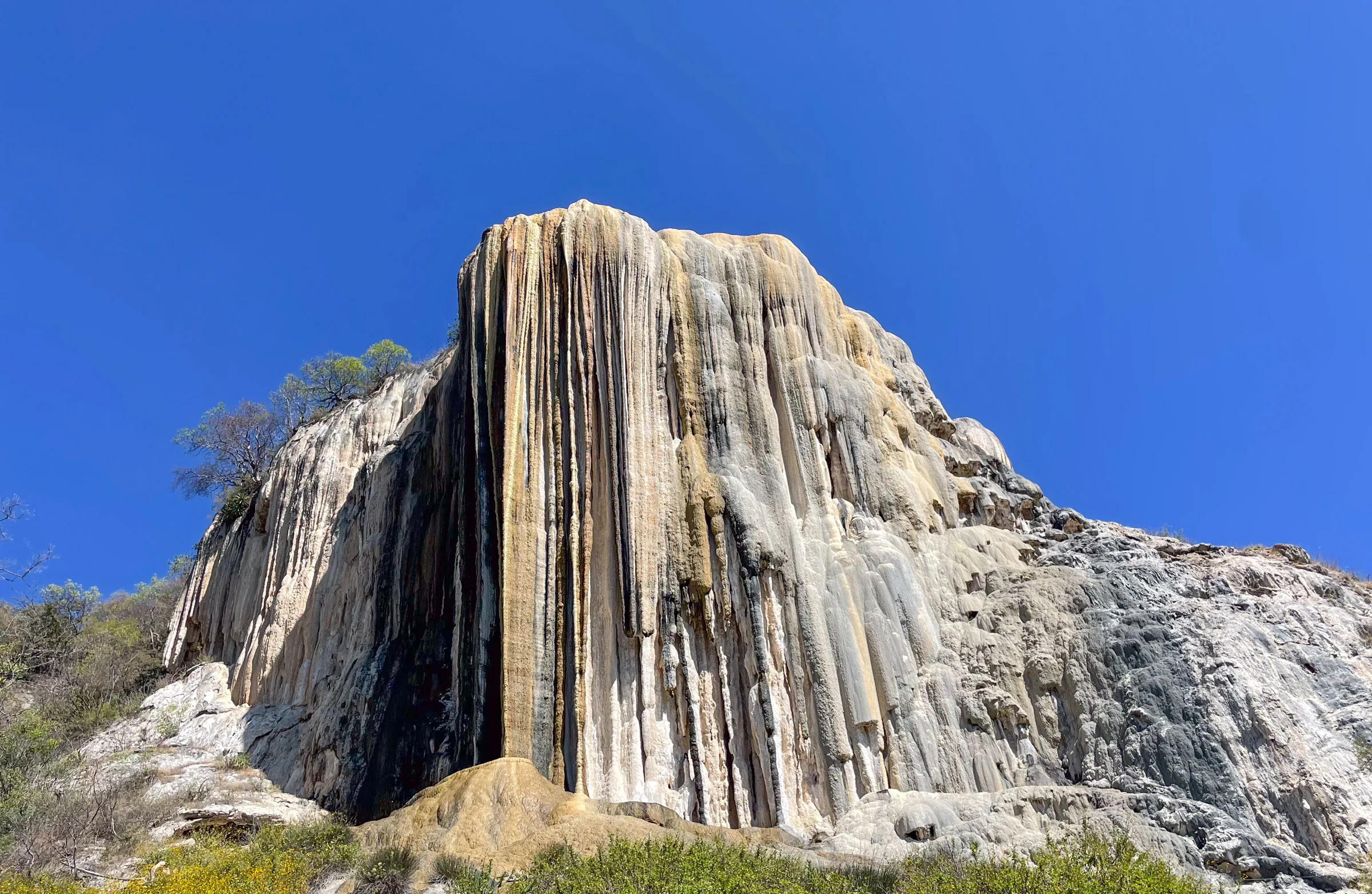 A tall cliff of white and ochre mineral deposits hanging in frozen streaks down a rock face, like a waterfall stopped mid-fall, under a deep blue sky