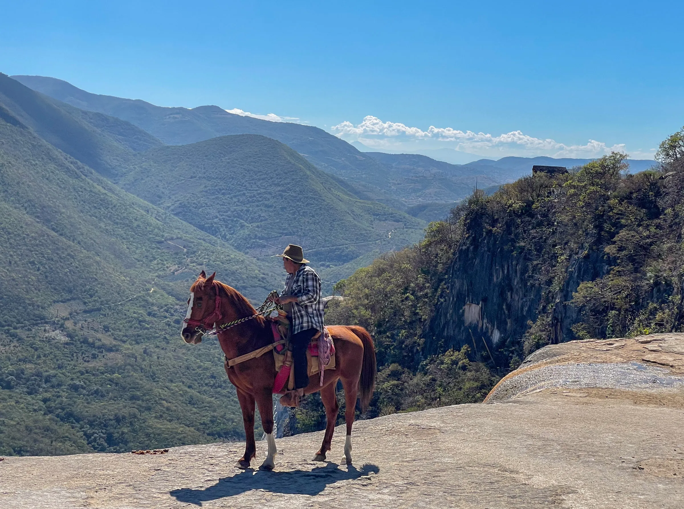 A rider in a straw hat and checked shirt sitting on a chestnut horse at the edge of a pale rock shelf, with a deep green valley and layered ridges behind