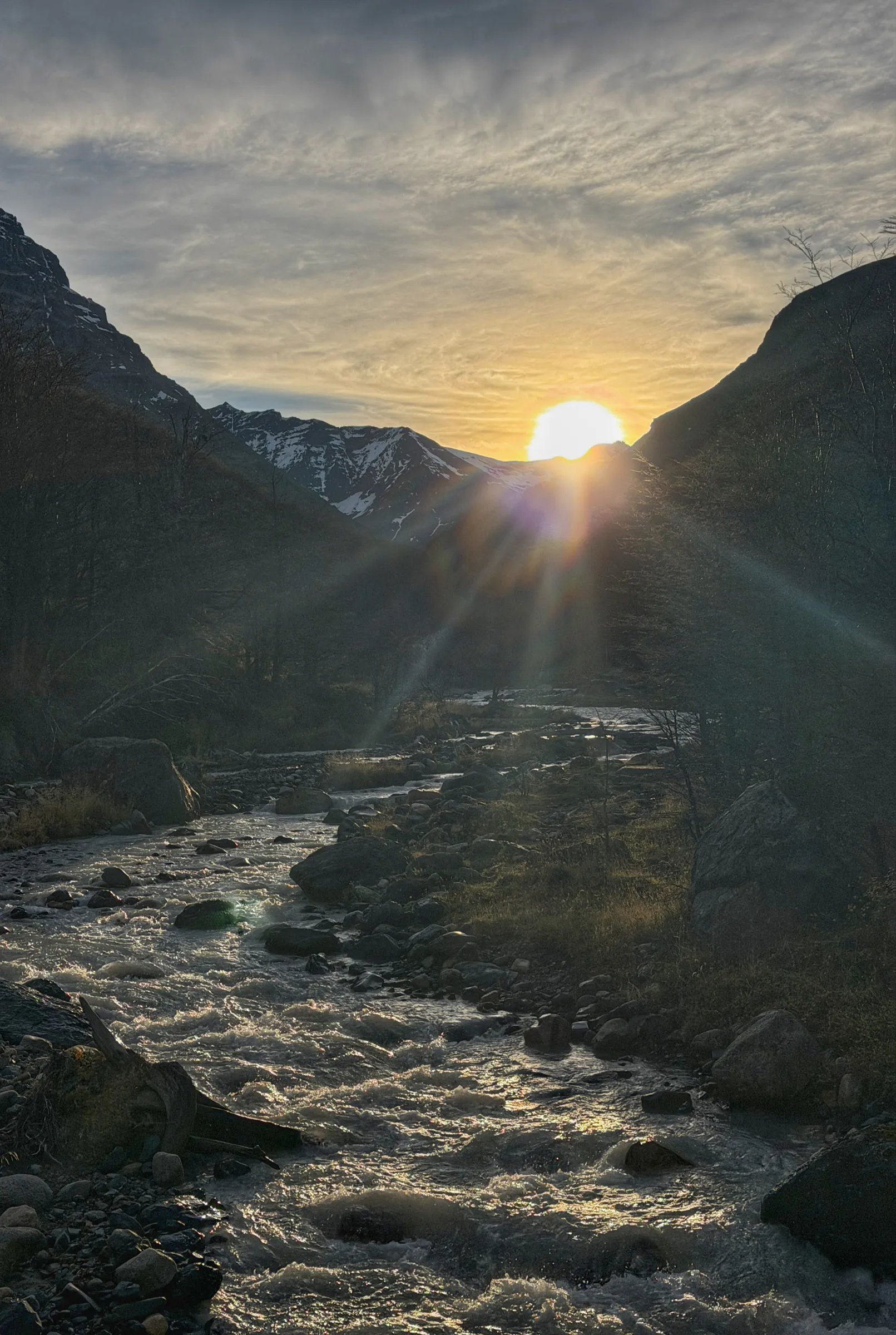 A shallow river running fast over boulders down a steep valley, with the sun setting into a notch between two dark ridges and snow on the peaks beyond