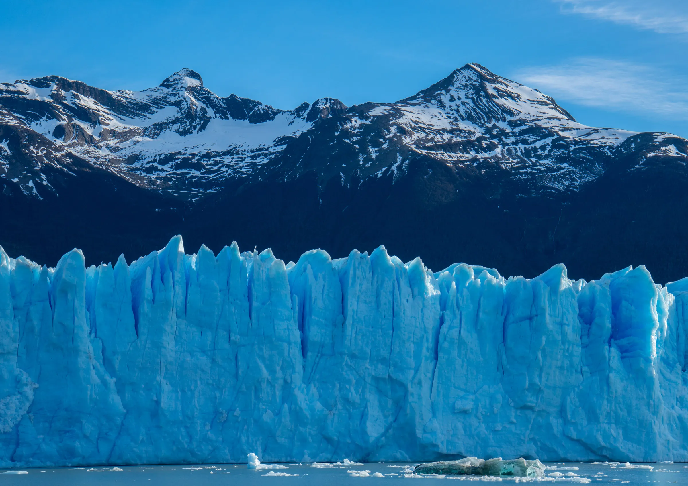The jagged blue ice wall at the front of a glacier meeting still grey water, with dark snow-streaked peaks behind