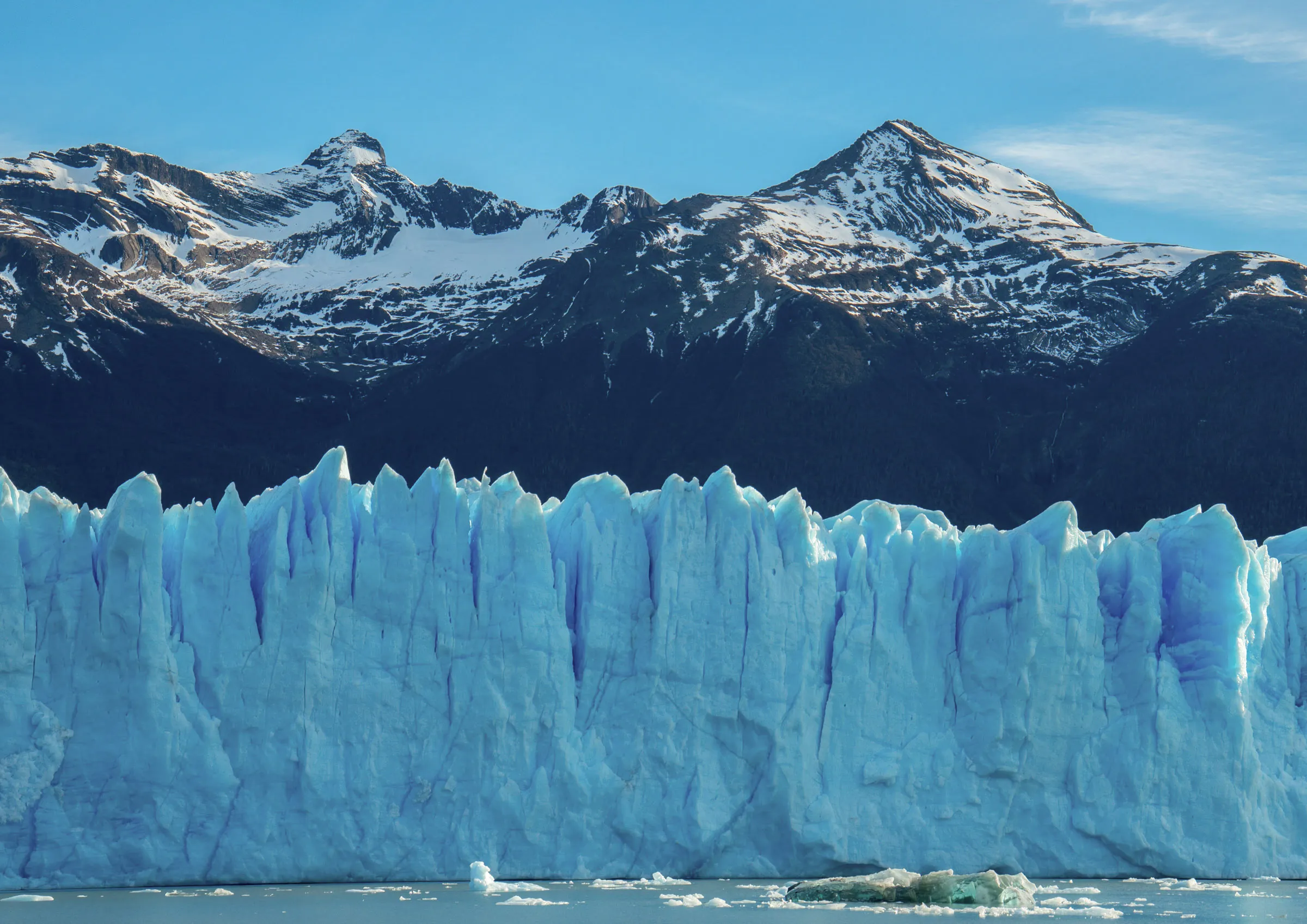 The jagged blue ice wall at the front of a glacier meeting still grey water, with dark snow-streaked peaks behind