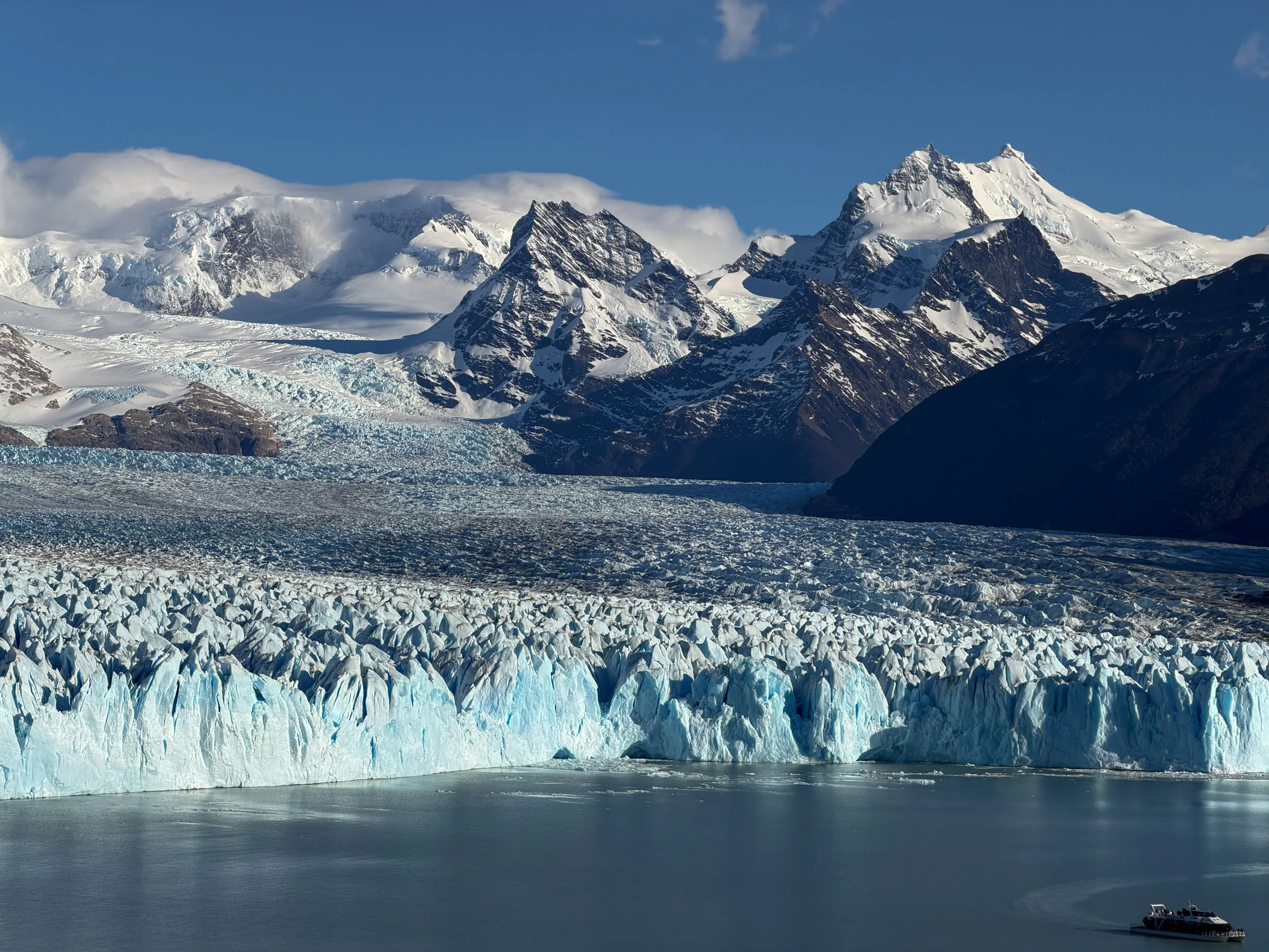 A wide blue-white glacier front meeting flat grey water, snow-covered peaks standing behind it and a small tour boat dwarfed against the ice