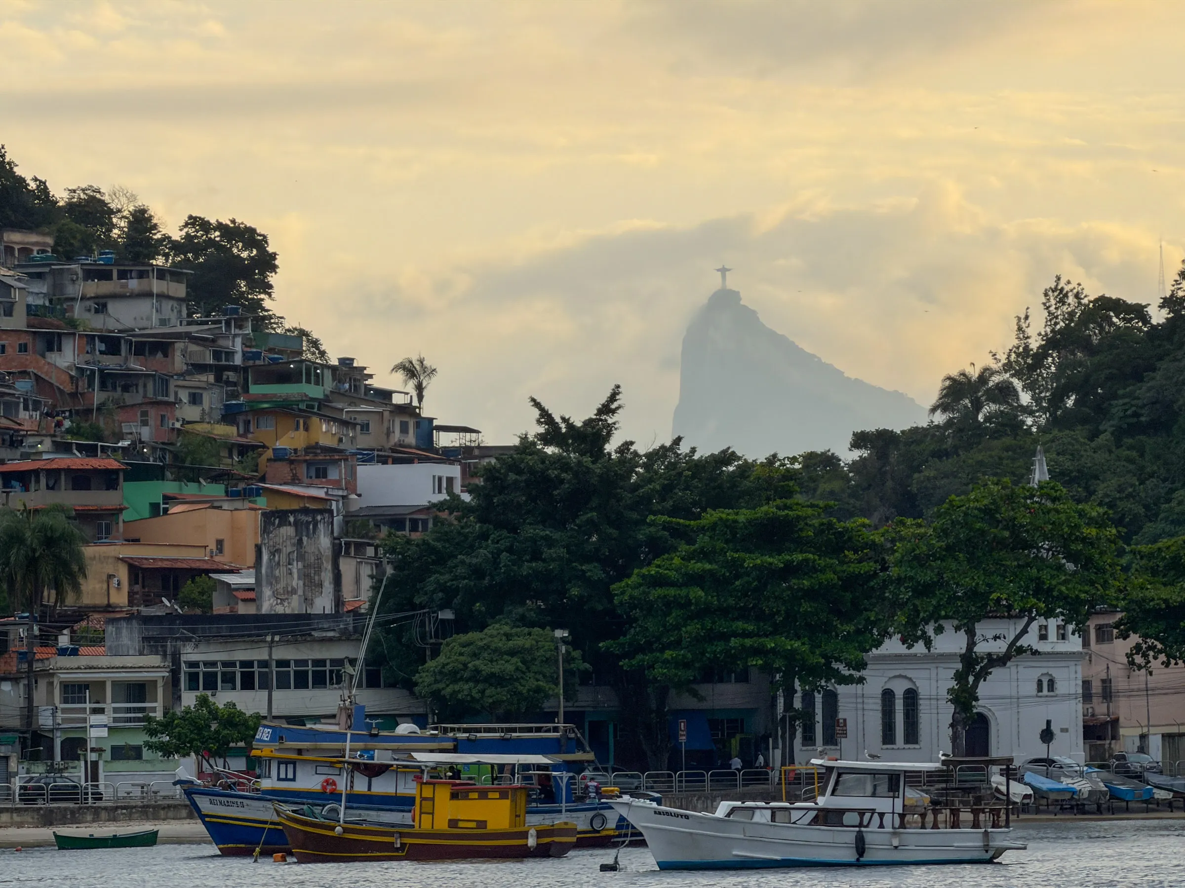 Fishing boats moored in front of a hillside of stacked houses at sunset, with the Christ the Redeemer statue small and hazy on the peak behind them