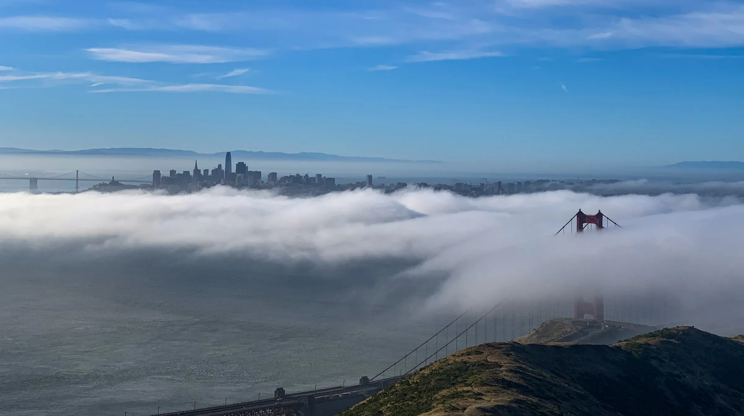 A low bank of fog filling the bay, with one tower of the Golden Gate Bridge rising through it and the San Francisco skyline on the horizon