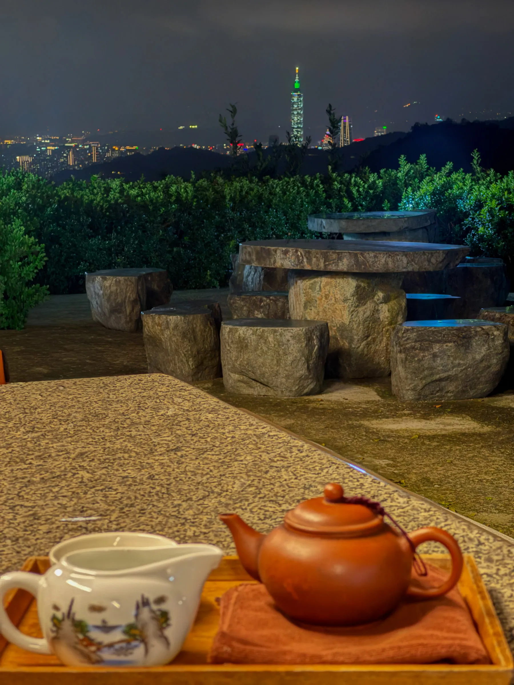 A clay teapot and white cups on a wooden tray in the foreground, with a stone table and stools on a dark terrace and the lit-up Taipei 101 tower on the skyline beyond