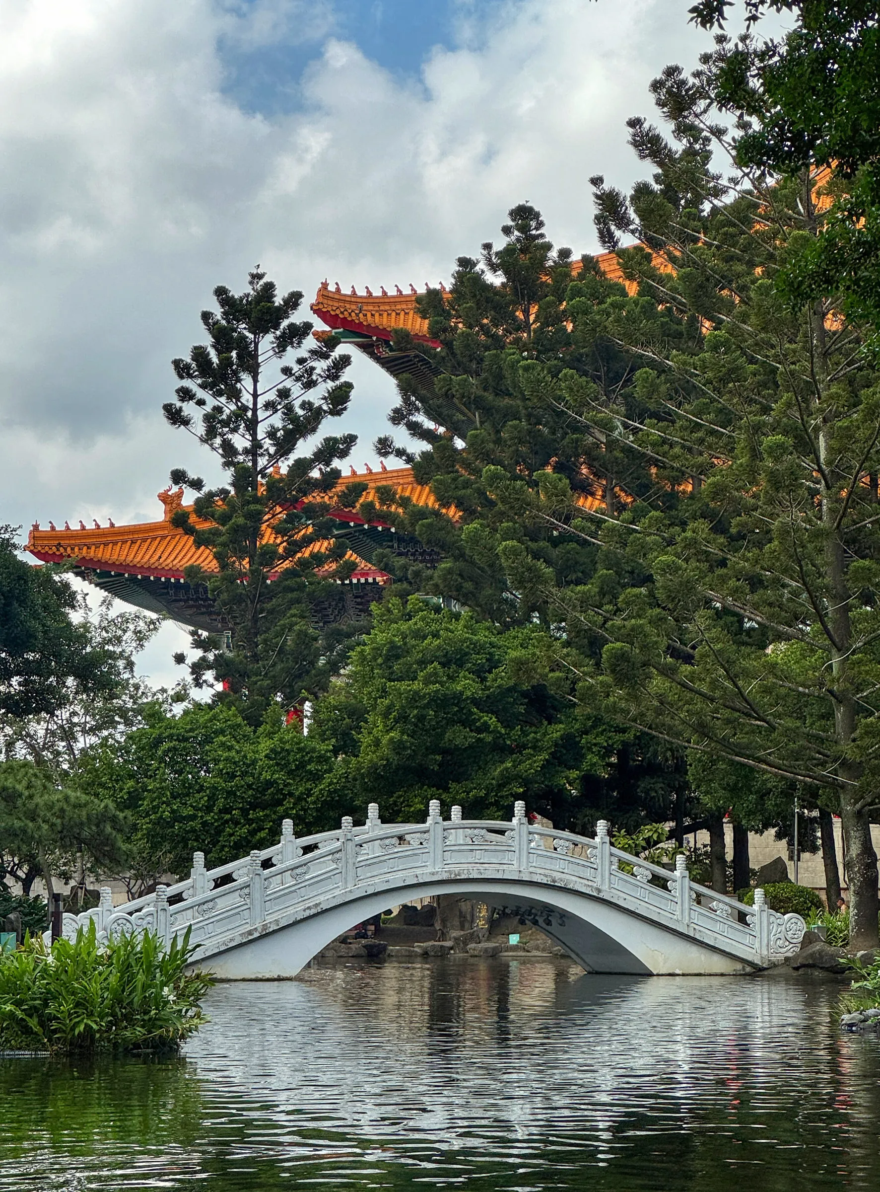 A white marble arched bridge over a park pond, with an orange-tiled temple roof rising above pine trees behind it