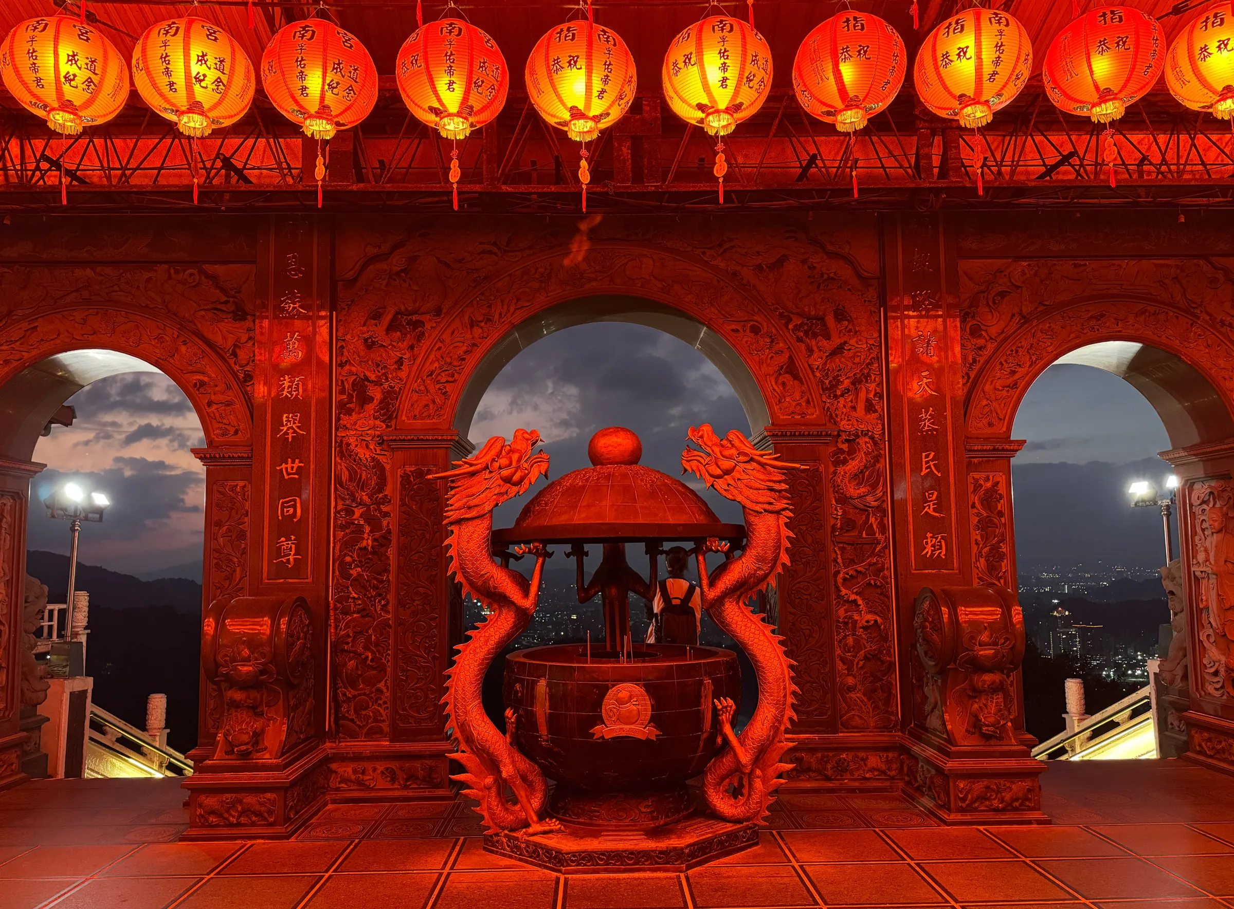 A red-lit temple archway with a twin-dragon incense burner in the foreground and rows of paper lanterns hanging overhead