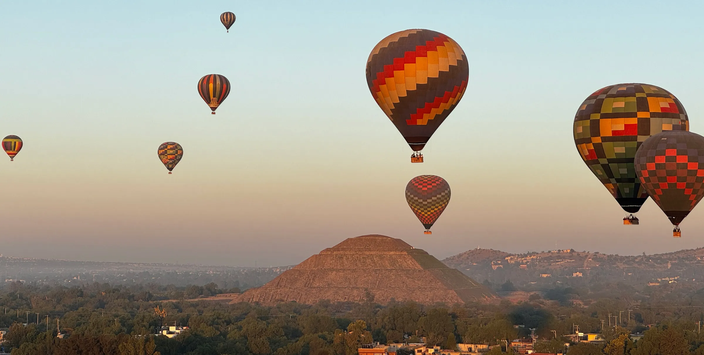 Hot air balloons drifting at sunrise above the Pyramid of the Sun, with the hazy valley spread out below