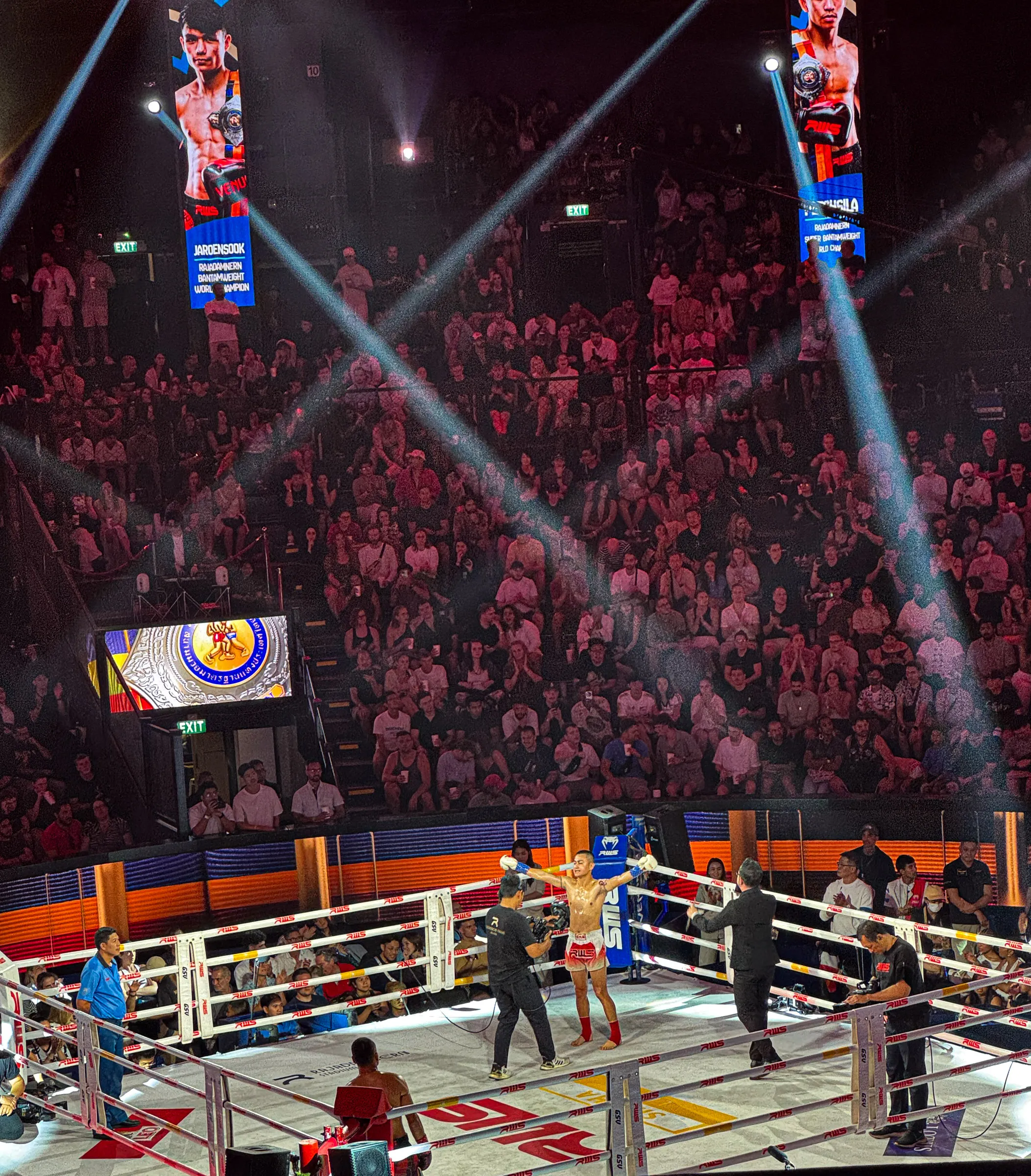 A Muay Thai fighter raising his arms in the ring at Rajadamnern Stadium, spotlights crossing overhead and a packed crowd filling the stands behind him