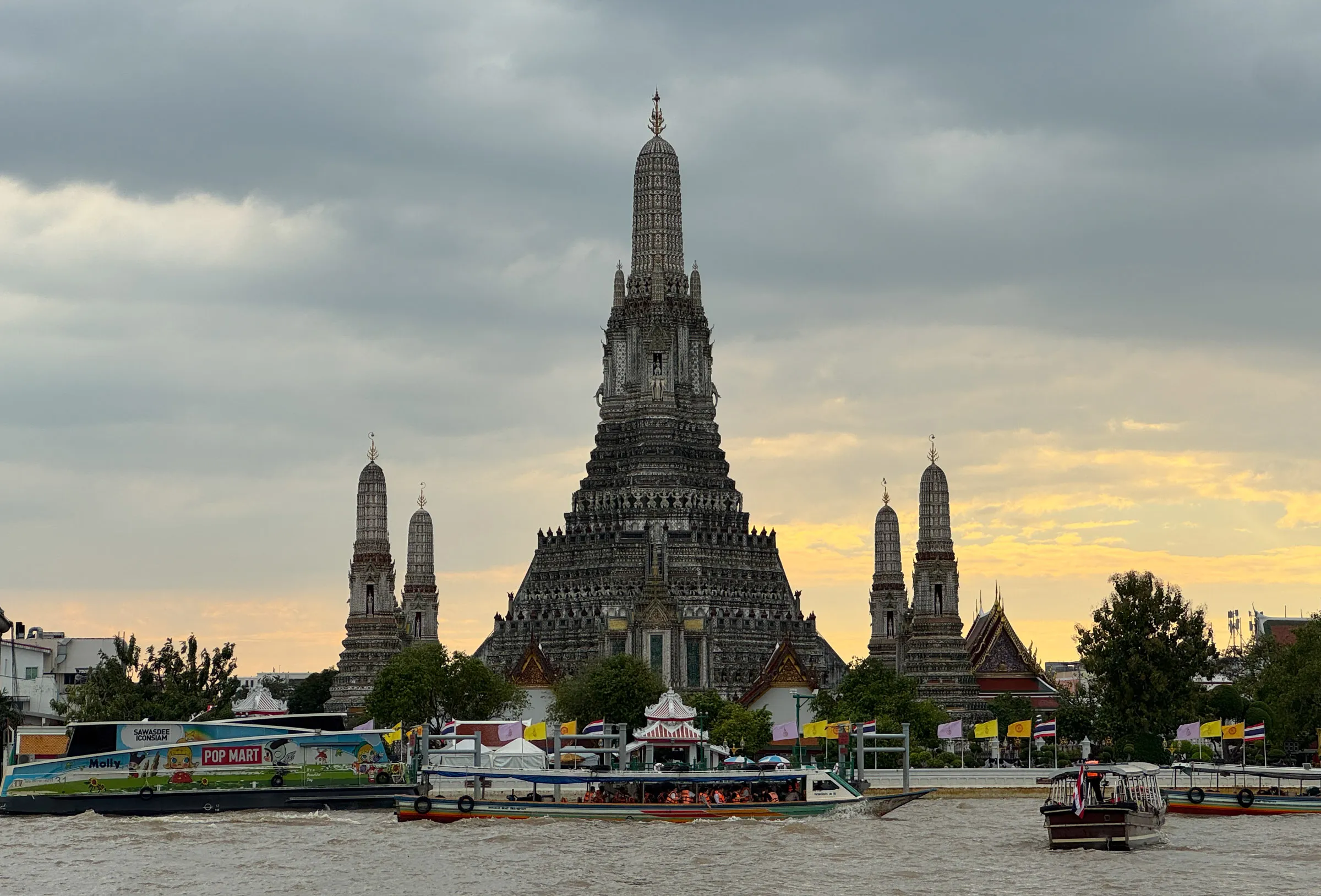 The tiered spires of Wat Arun rising above the Chao Phraya River at dusk, with boats crossing the water in front of it