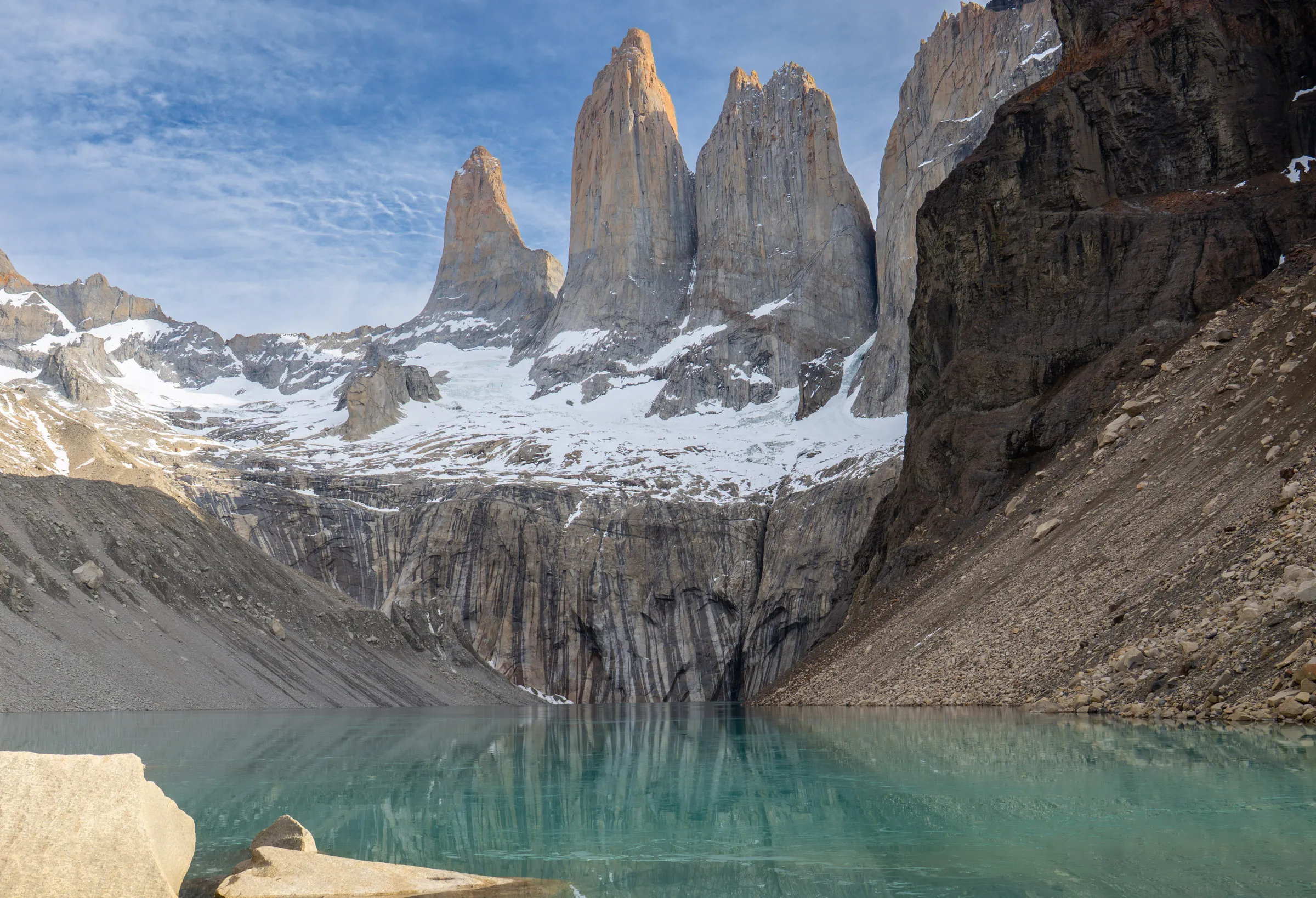 Three granite towers rising above a snowfield and a turquoise glacial lake under a pale blue sky